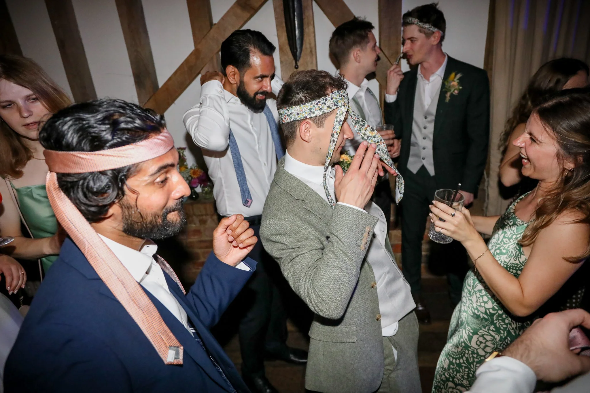 People at a celebration, some wearing colorful headbands and casual attire, socializing and smiling in a rustic wooden setting.