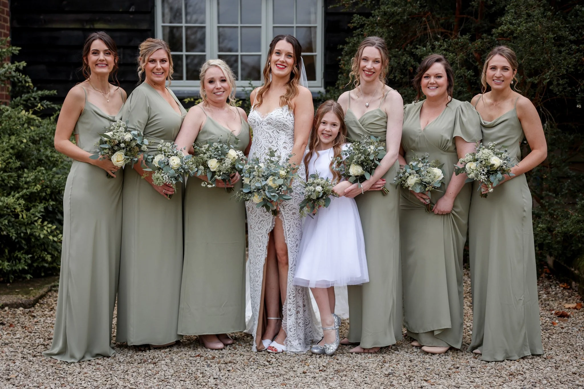 A group of nine women, including a young girl, standing outdoors in front of a black building with a large window, all dressed in formal attire for a wedding or special occasion, holding bouquets of white and green flowers.