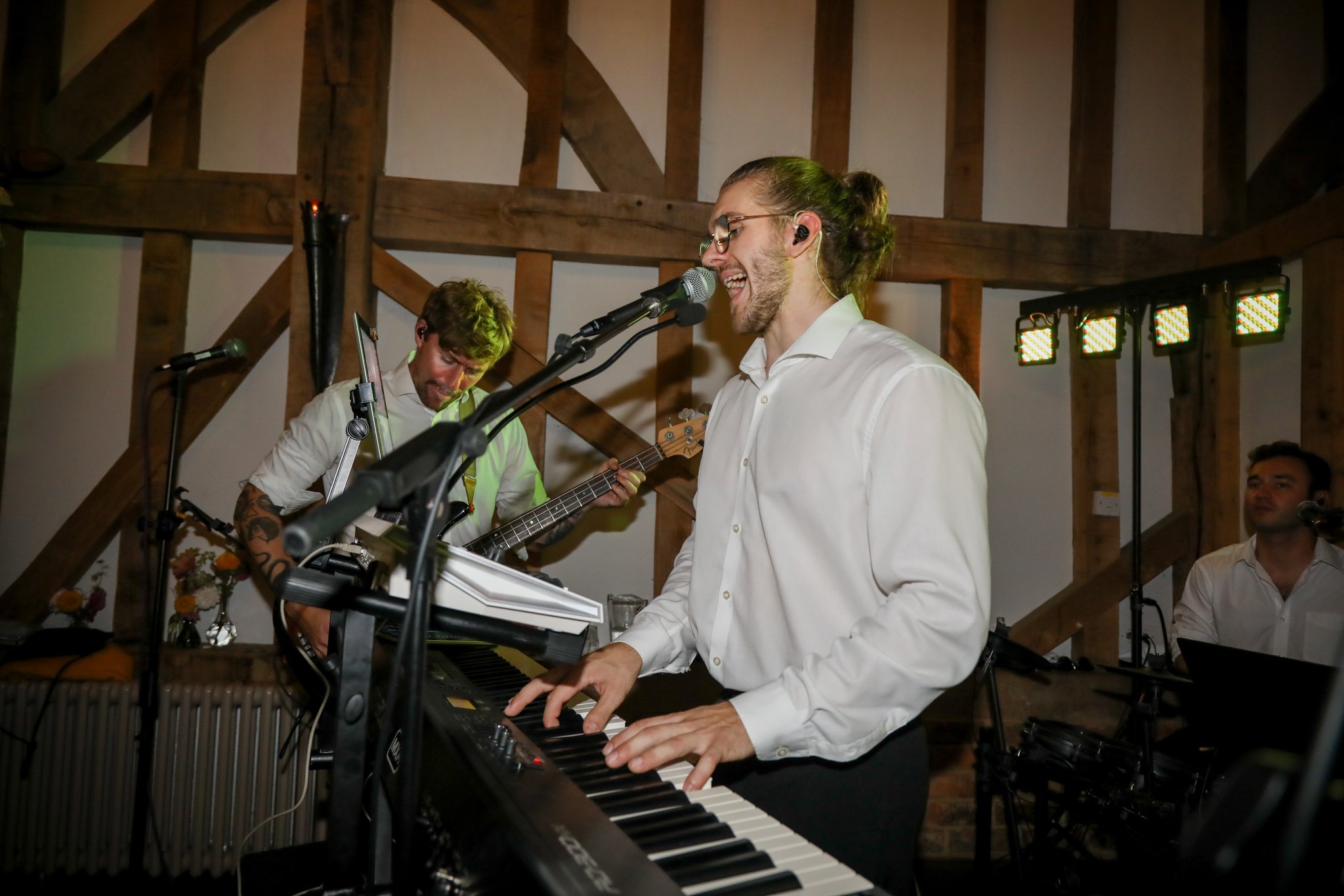 A musician plays keyboard and sings into a microphone at an indoor event with wooden beams and colorful stage lights.