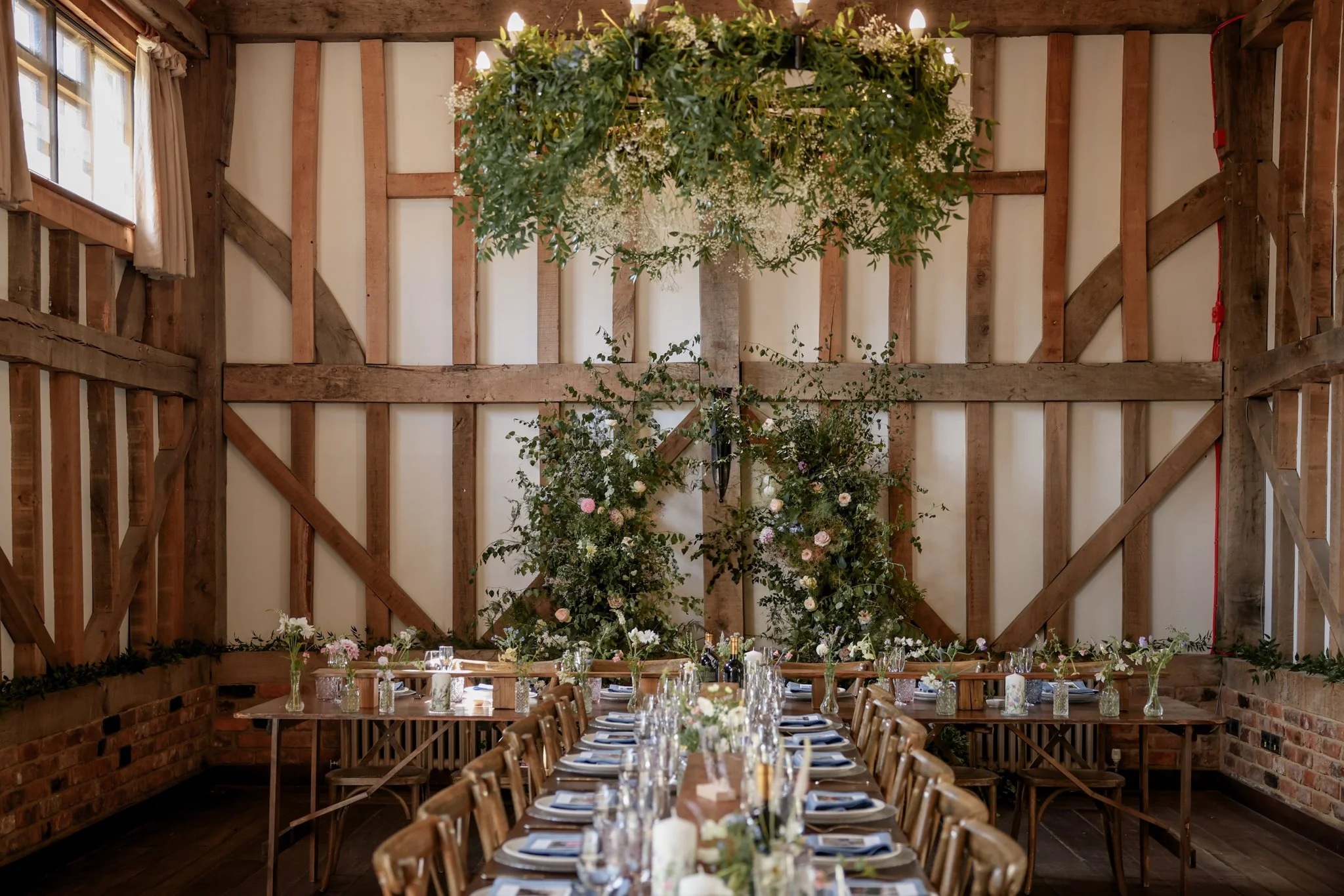 Wedding reception setup in a rustic venue with long tables decorated with flowers, glasses, and plates, and a large floral chandelier hanging from a wooden ceiling.