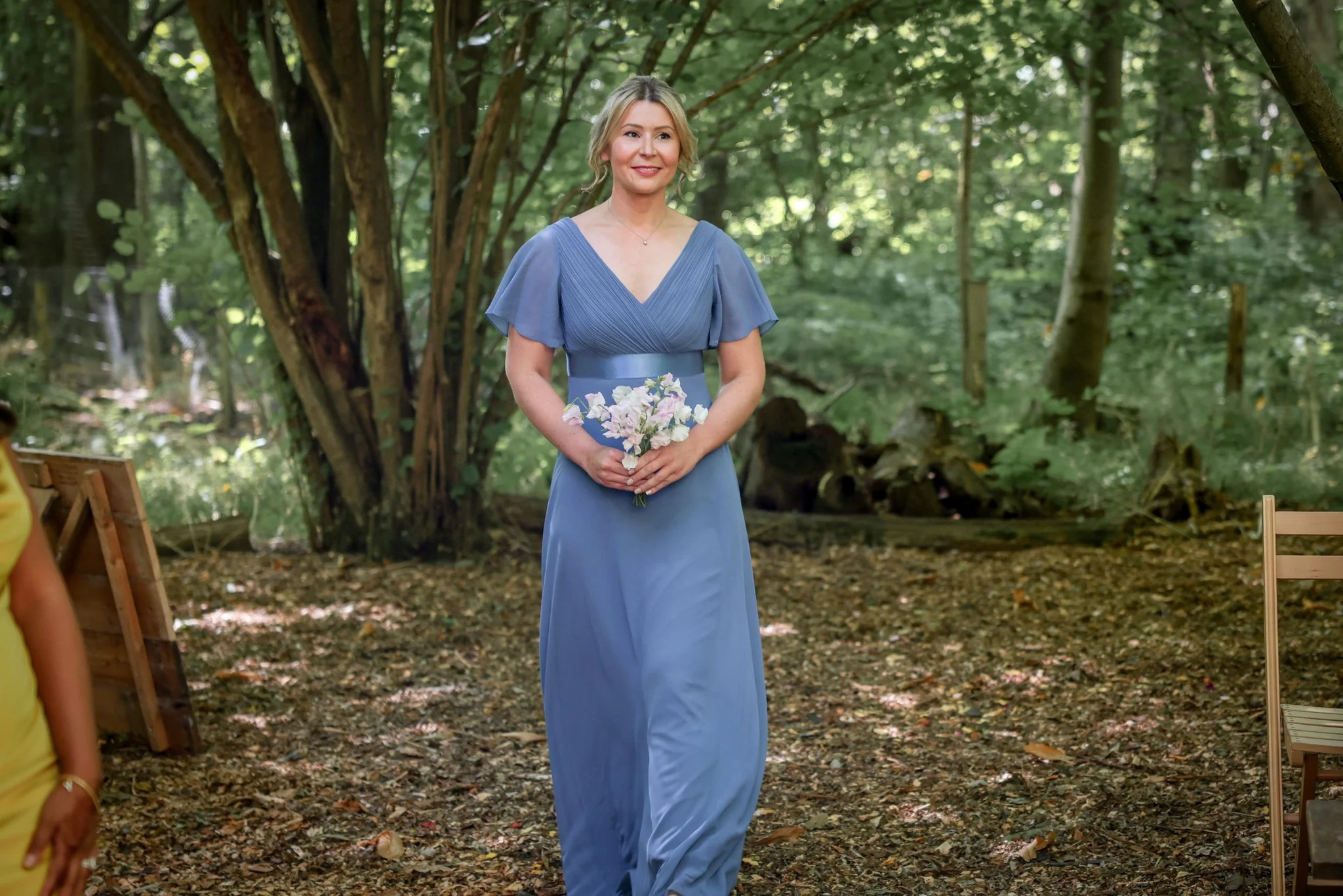 A woman in a blue dress holding a small bouquet of flowers standing in a wooded outdoor setting.