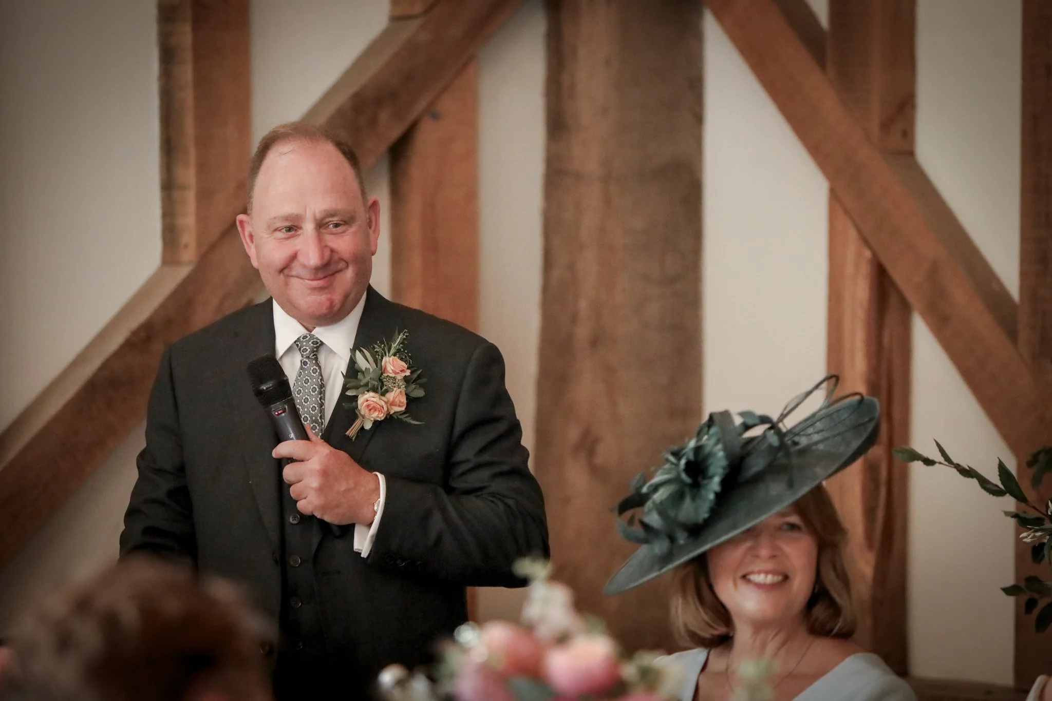 A man in a black suit holding a microphone and smiling, with a woman in a white dress and floral fascinator, also smiling, in a wooden-beamed room at what appears to be a wedding celebration.