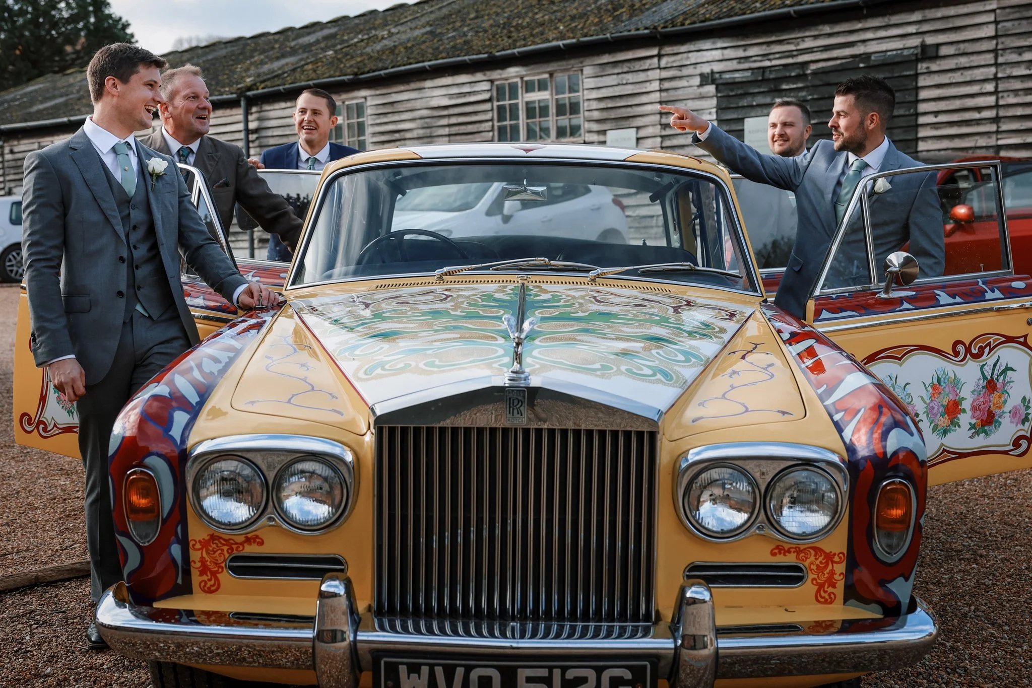Groom and groomsmen in suits with boutonnieres standing around a vintage yellow Rolls Royce decorated with colorful floral patterns, outdoors near a rustic wooden building.