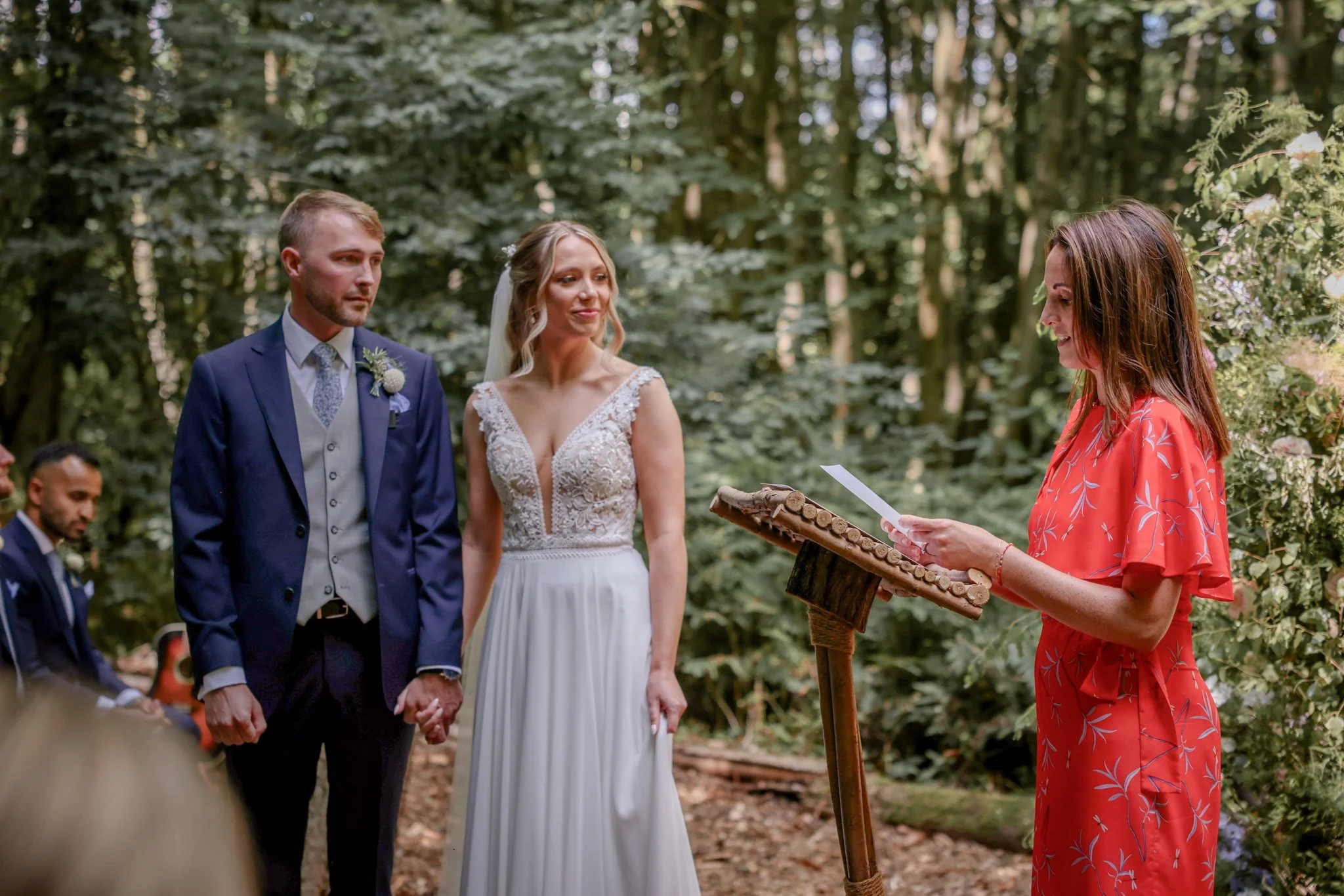 A couple gets married outdoors in a wooded area during the daytime, holding hands and standing before a woman officiating their wedding ceremony.