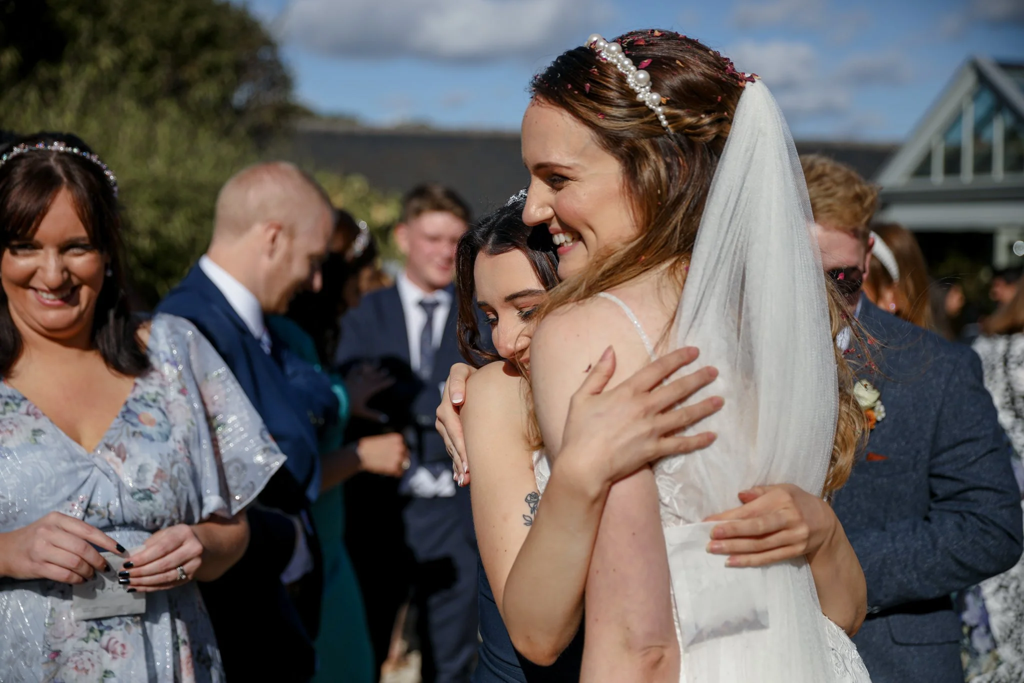 A bride hugging a woman during a wedding celebration outdoors with a crowd of guests in the background.
