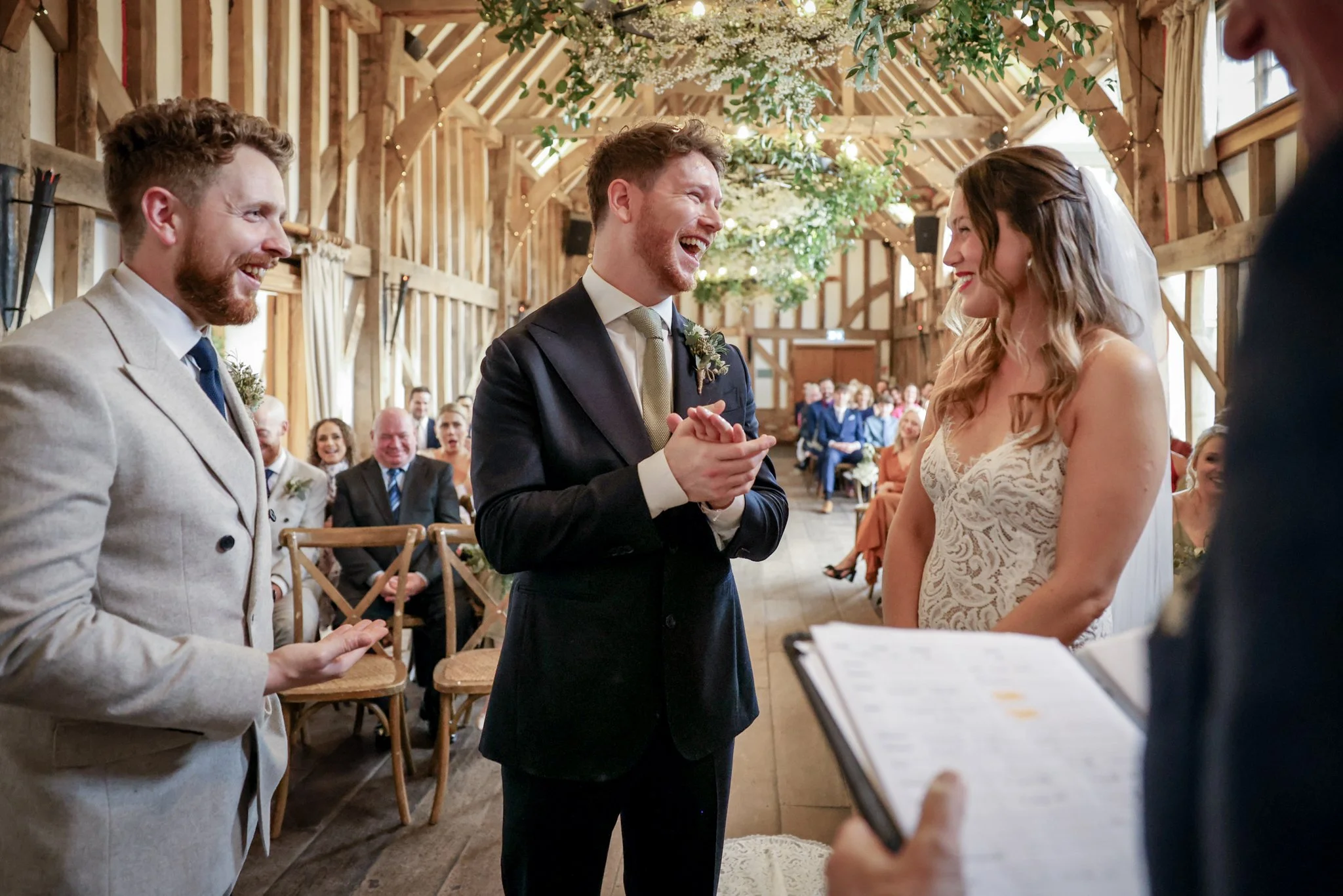 A groom and bride exchanging vows during their wedding ceremony inside a rustic barn with guests seated in the background. The groom is smiling and facing the bride, while the bride is also smiling and facing the groom. An officiant is partially visi