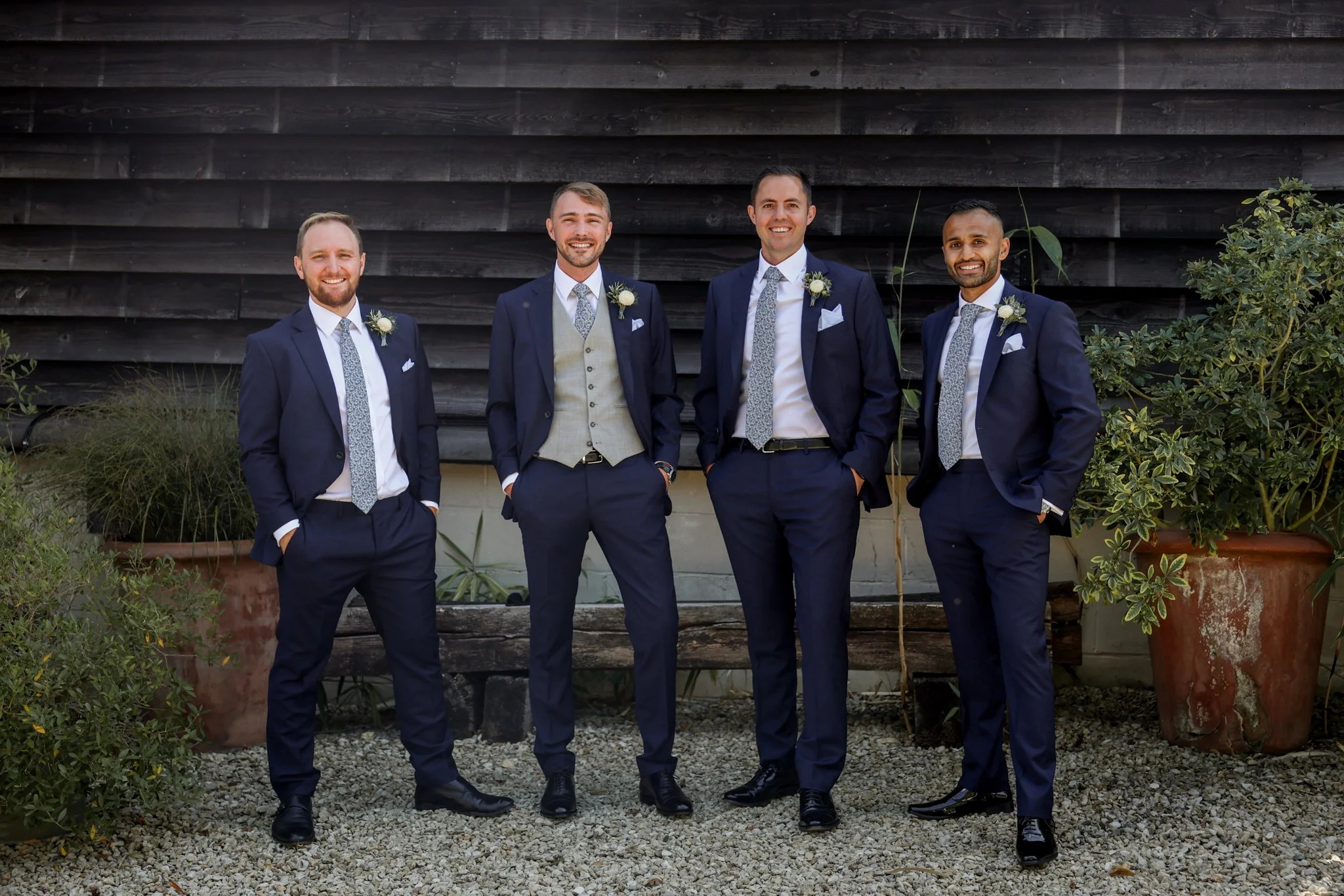 Four men dressed in formal suits, standing outdoors against a wooden wall, smiling for the photo with some greenery around them.