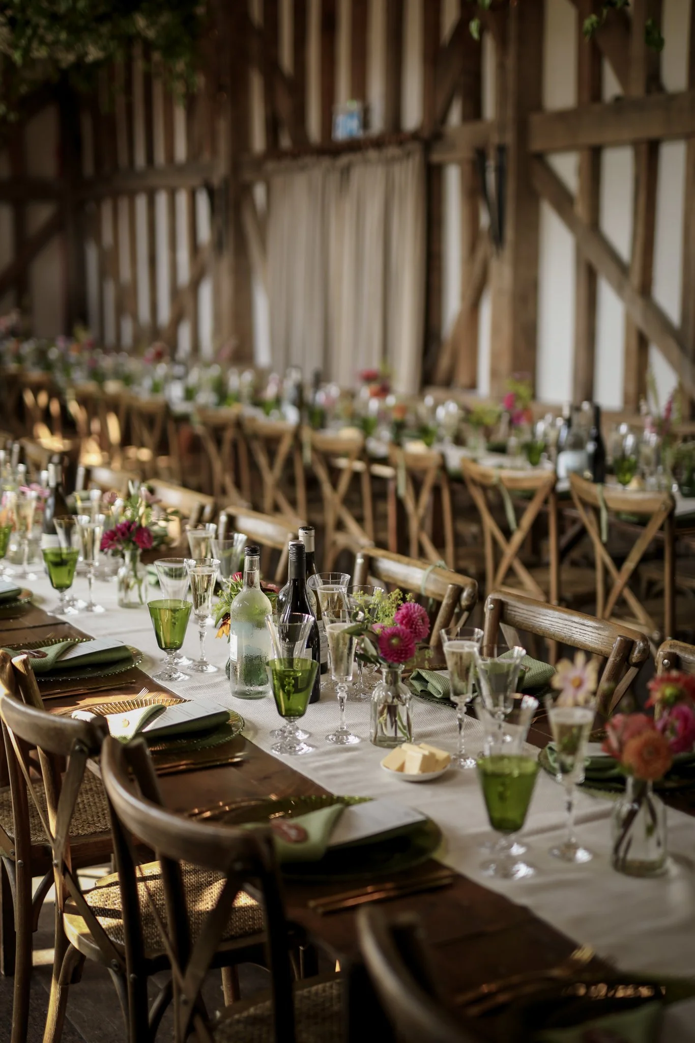 A rustic indoor dining setup featuring a long wooden table with a white tablecloth, decorated with pink and green floral centerpieces, glassware, and dinner settings, set against a backdrop of wooden barn walls and beams.