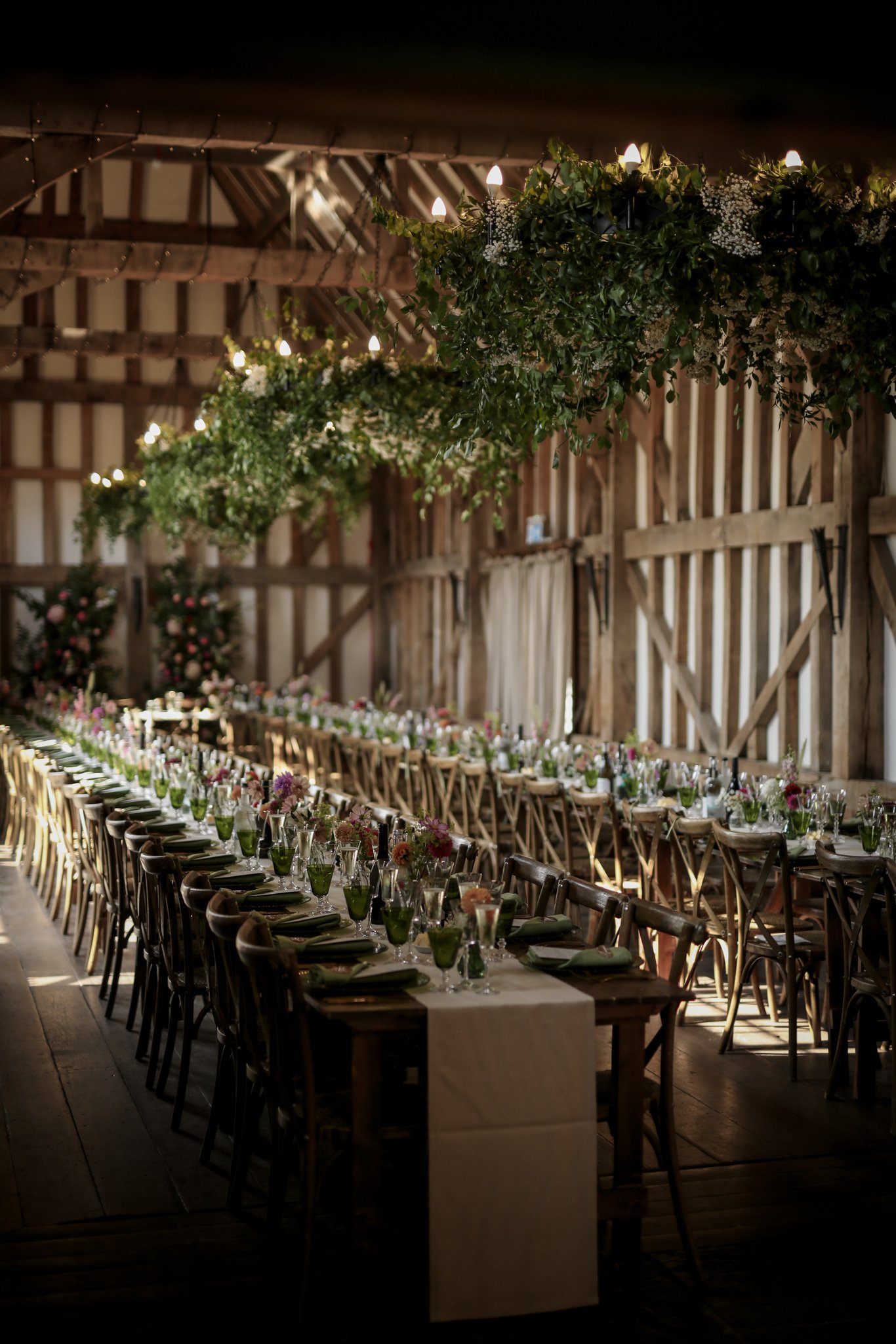 Long banquet table decorated with flowers and green glasses, set up in a rustic barn with wooden walls and hanging greenery and lights.