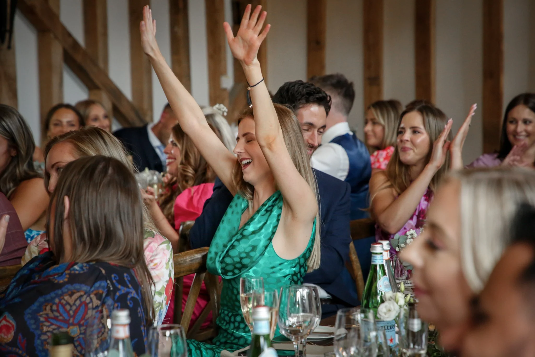People celebrating at a social gathering, some with their hands raised and smiling, with drinks and bottles on the table, in a rustic indoor setting with wooden beams.