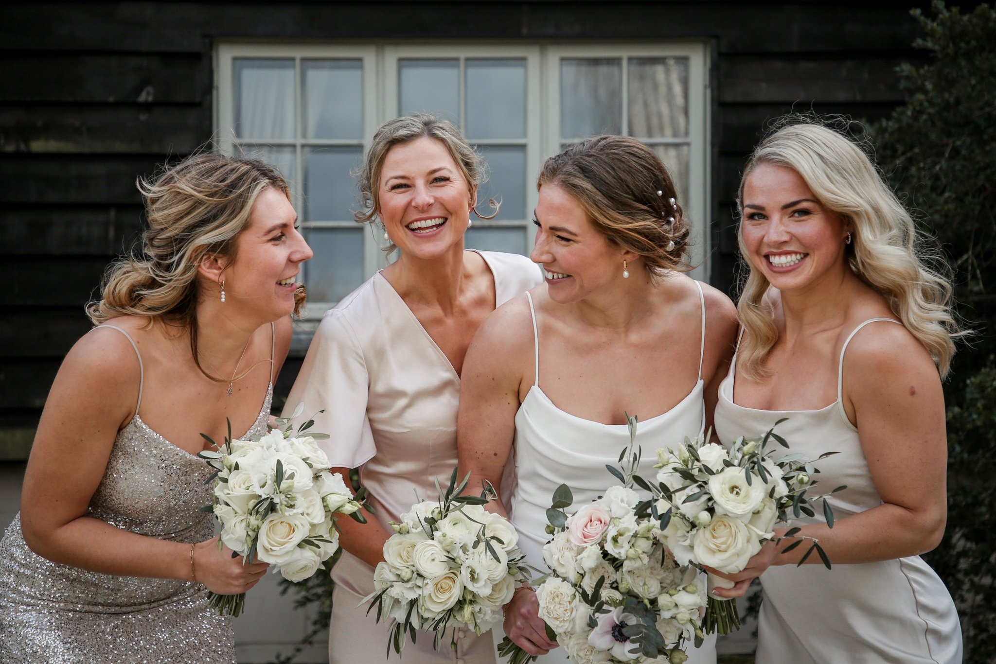 Four women in wedding dresses holding bouquets of white flowers, smiling and looking at each other outside of a dark building with a large window.