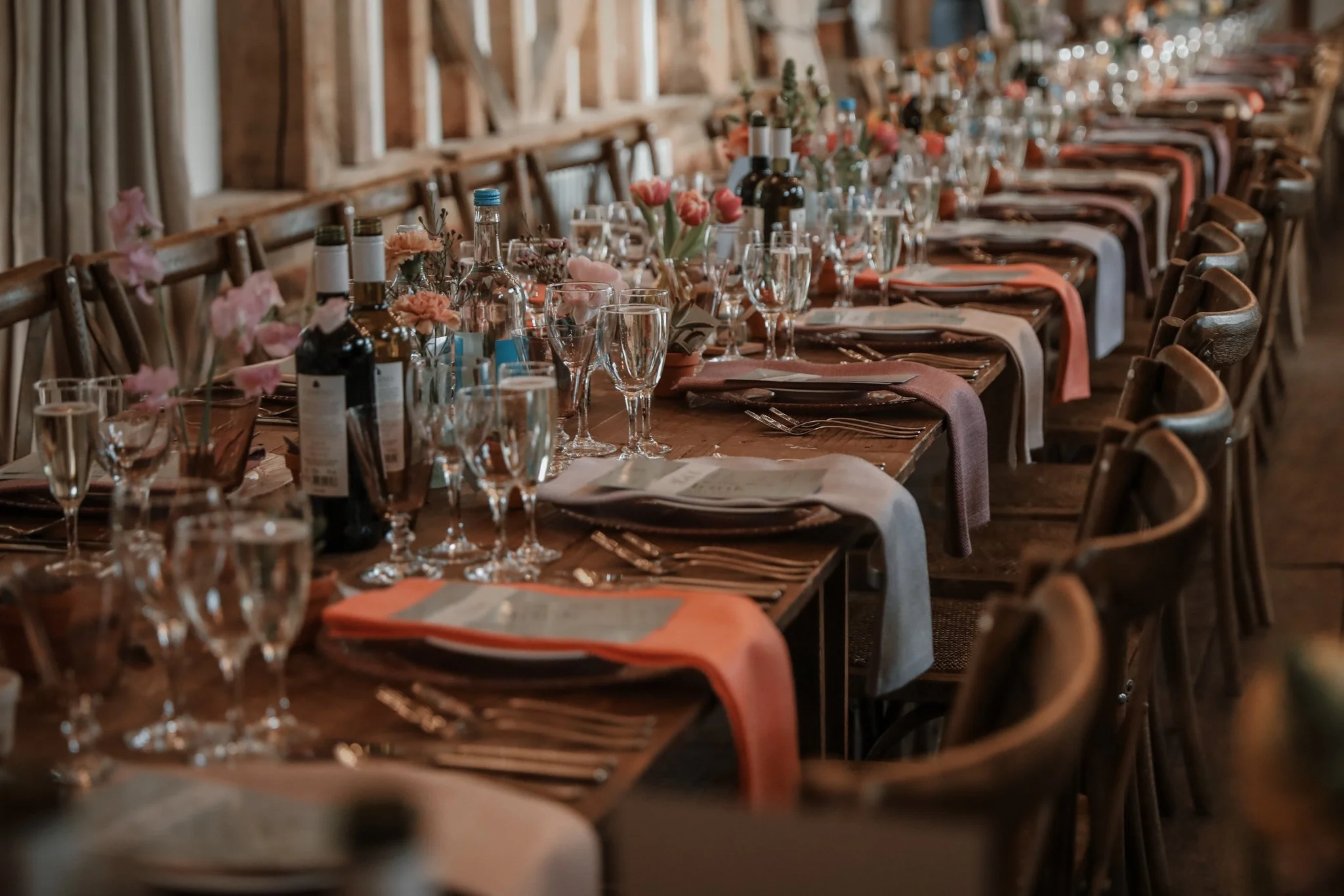 Long banquet table set for a celebration with pink flowers, wine bottles, glasses, and neatly folded napkins, in a rustic wooden venue.