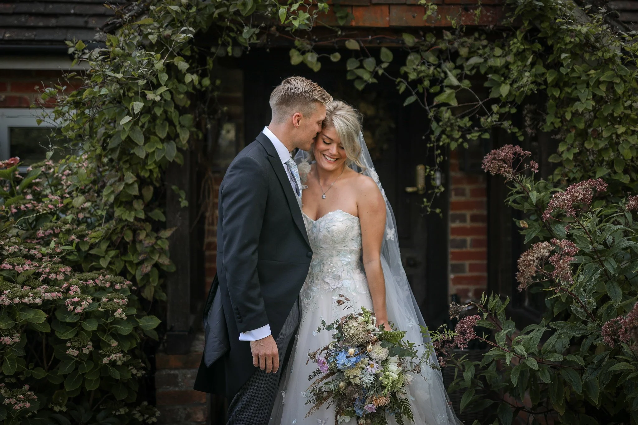 A newlywed couple is standing close together, smiling, and touching foreheads outdoors behind greenery and a brick wall.