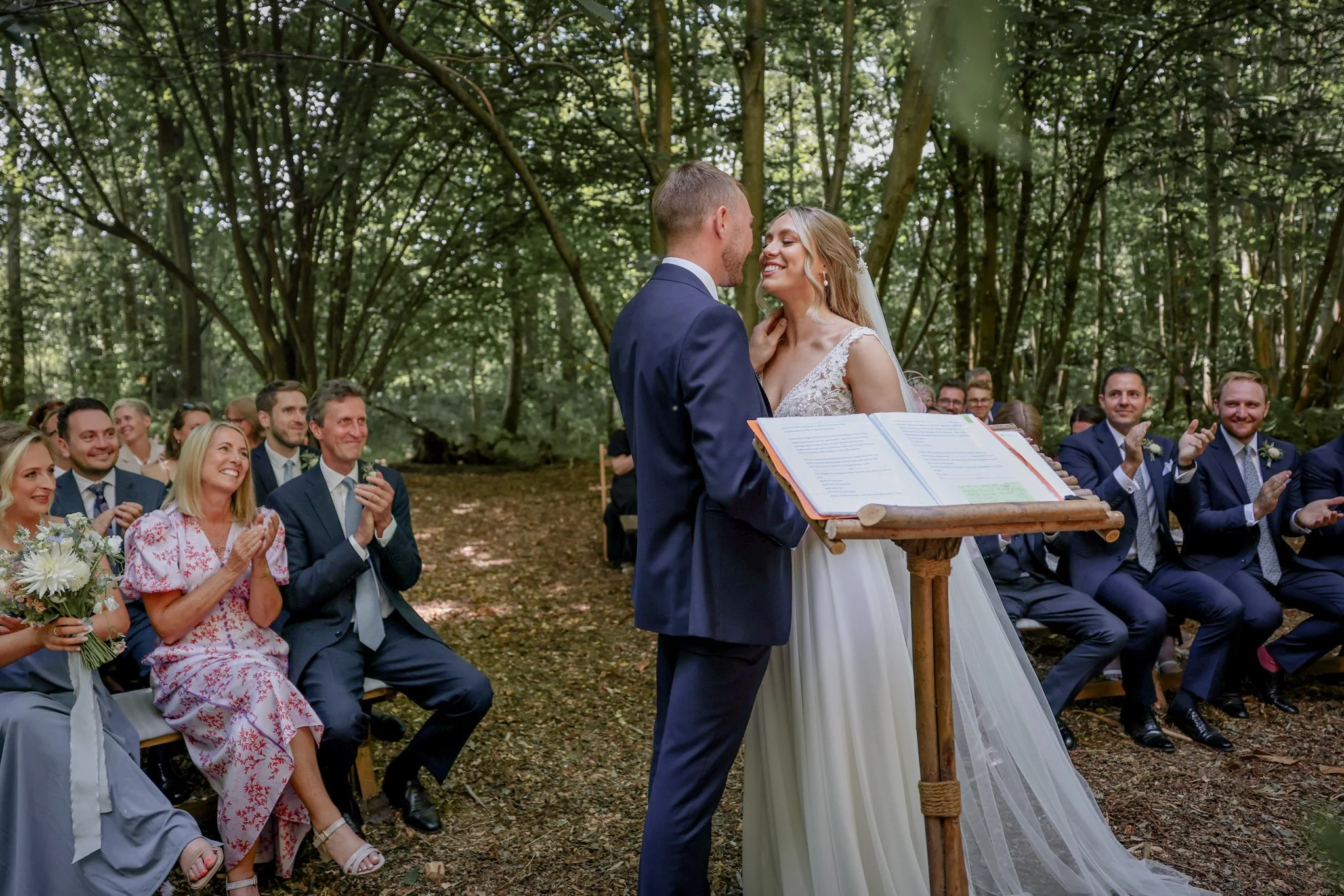A bride and groom exchange vows during an outdoor wedding ceremony in a wooded area, with seated guests applauding.
