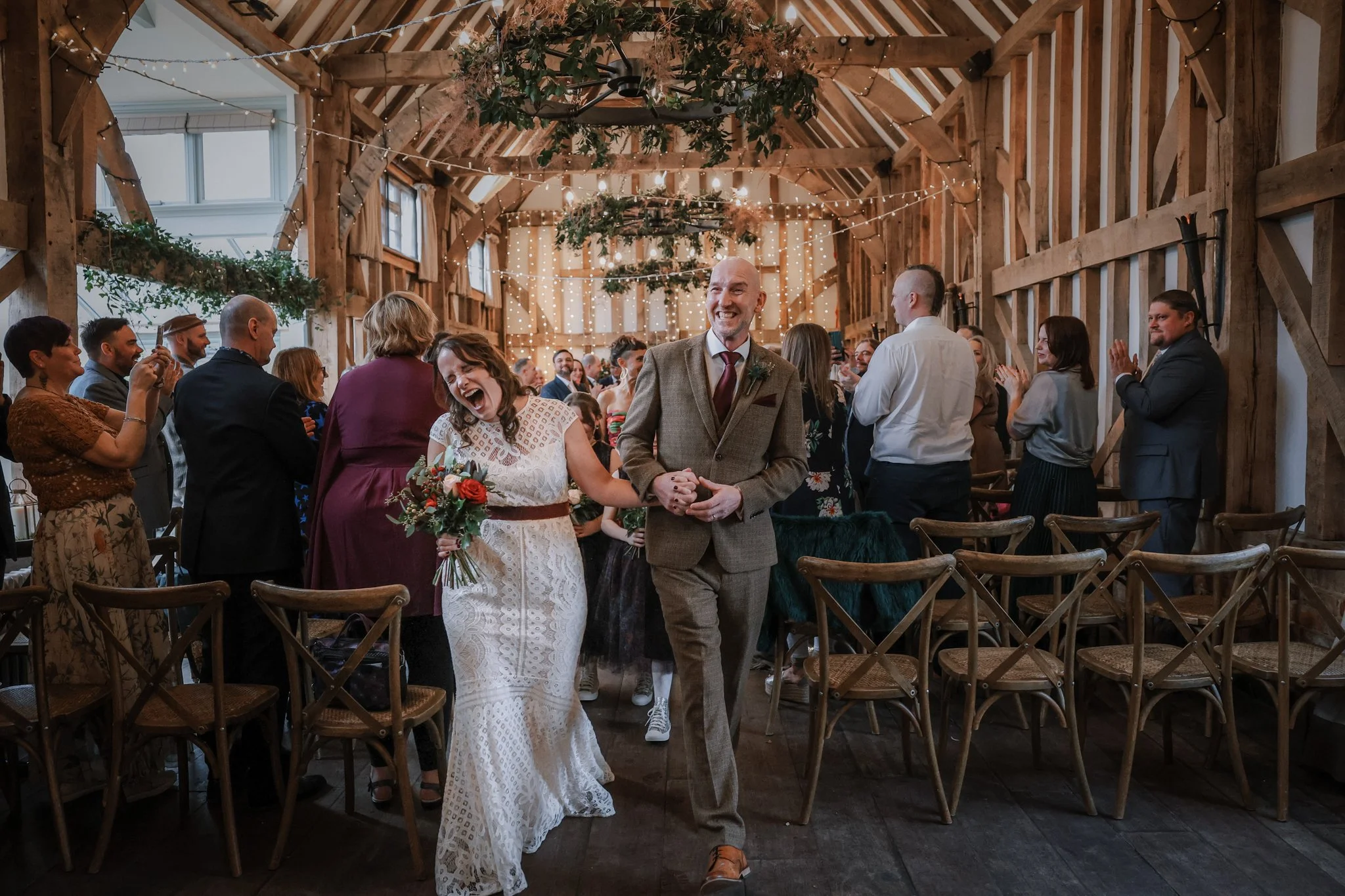 A bride and groom are walking together down the aisle at their wedding ceremony inside a rustic venue decorated with string lights and hanging greenery, with guests standing and smiling around them.