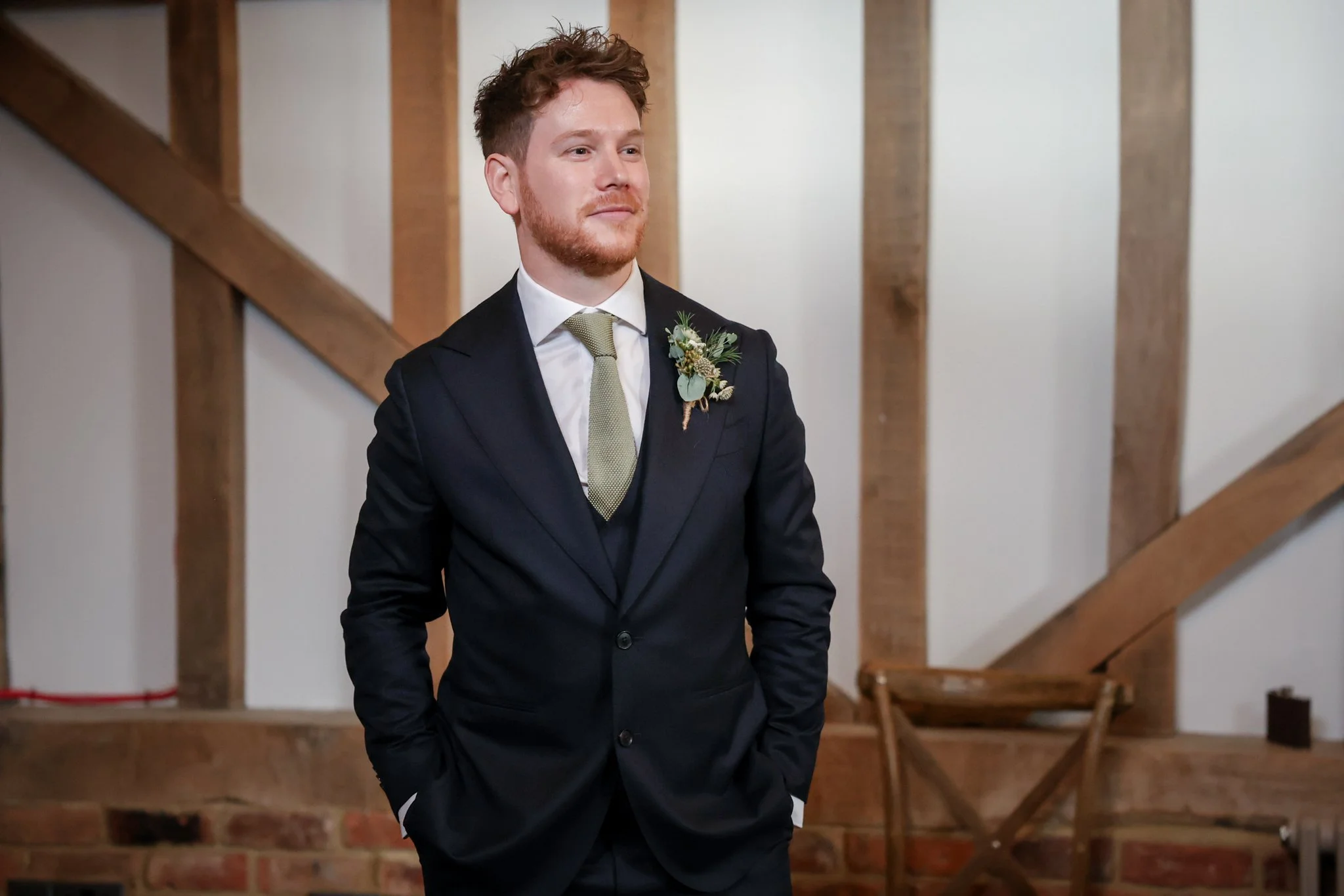 A young man with reddish hair and a beard wearing a dark suit with a white shirt and a light green tie, standing indoors in front of a wooden staircase with a serious expression.