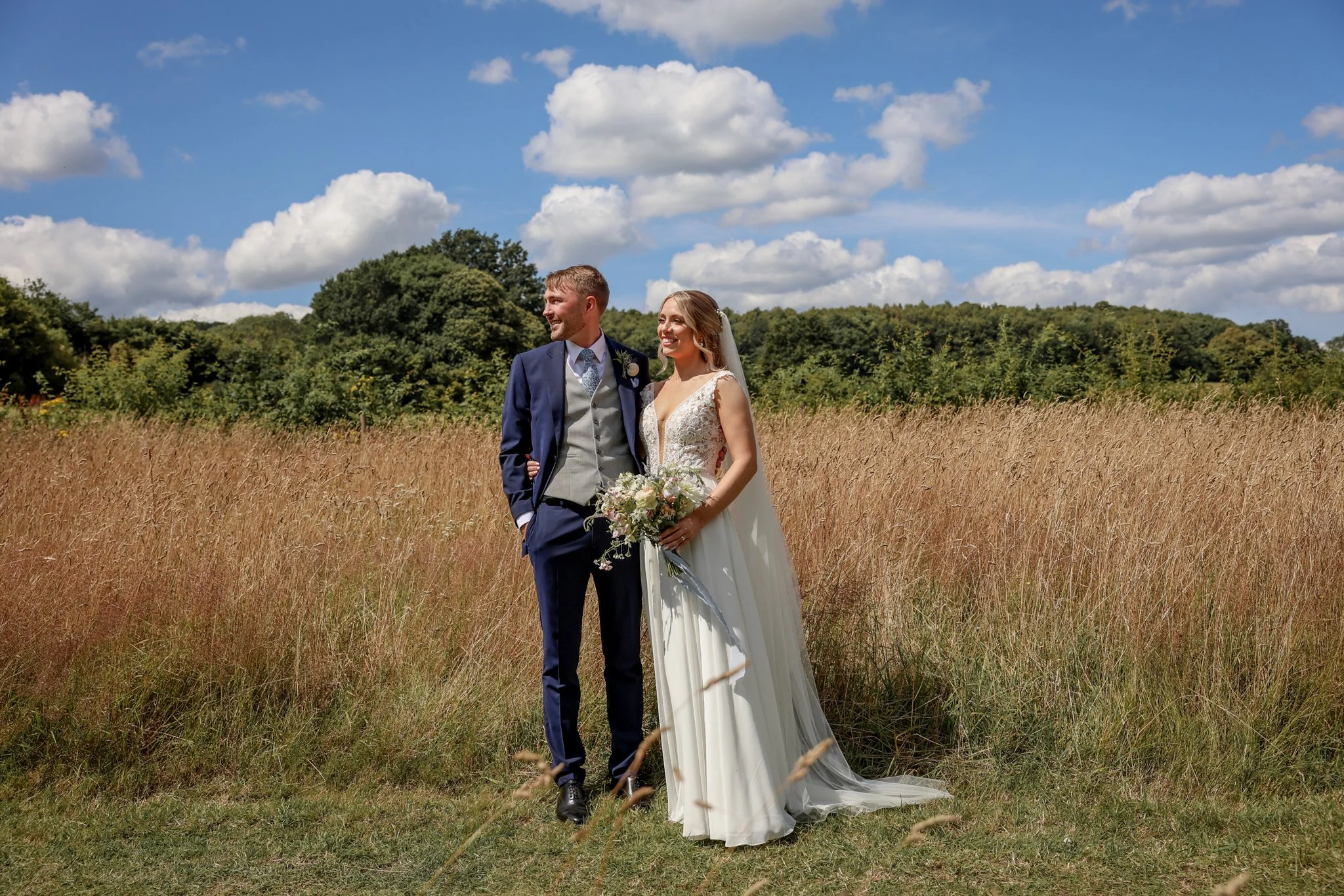 A bride and groom, both smiling, standing in a field of tall grass with trees and a partly cloudy sky in the background.