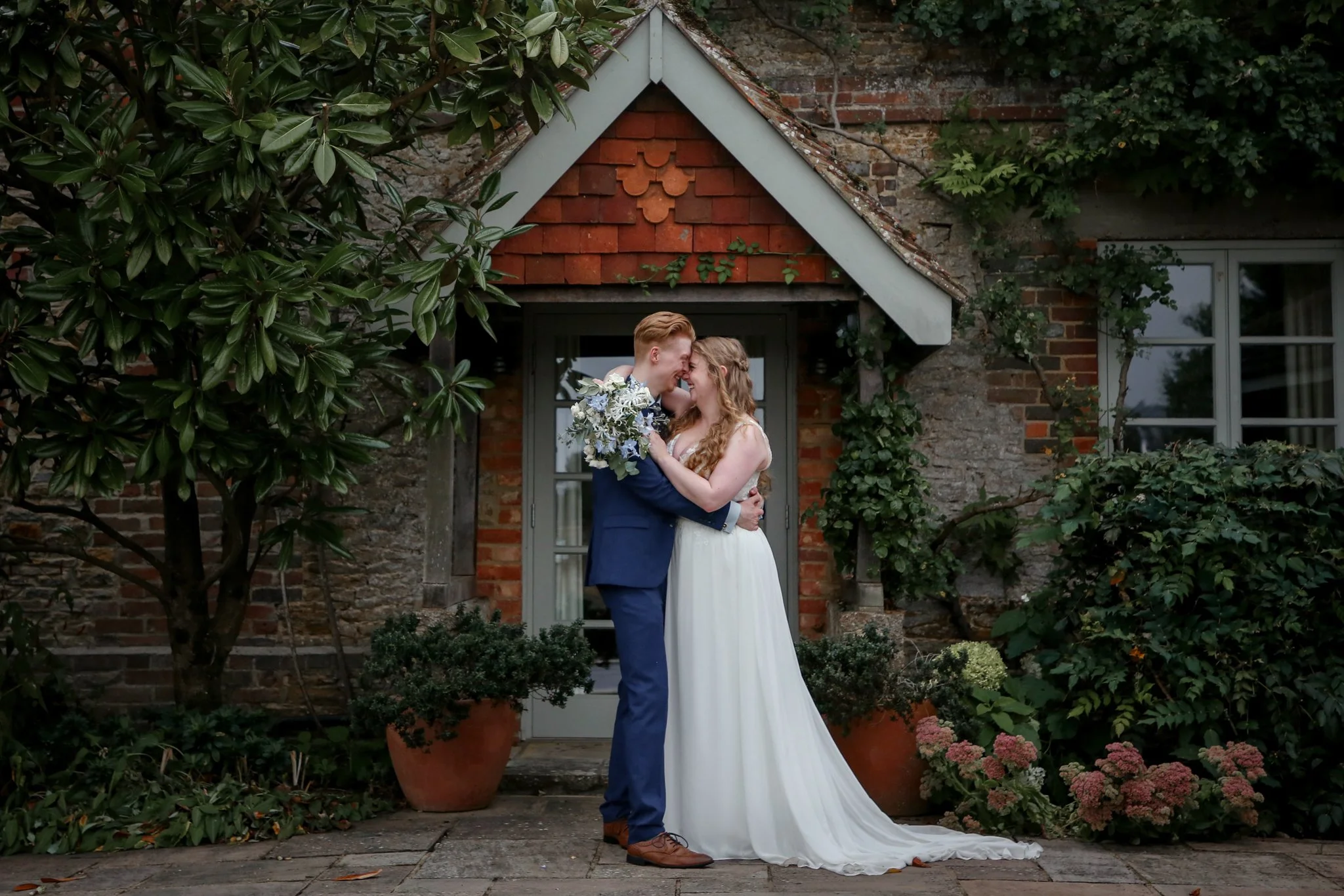 A bride and groom sharing a kiss outside a brick house with greenery around them.