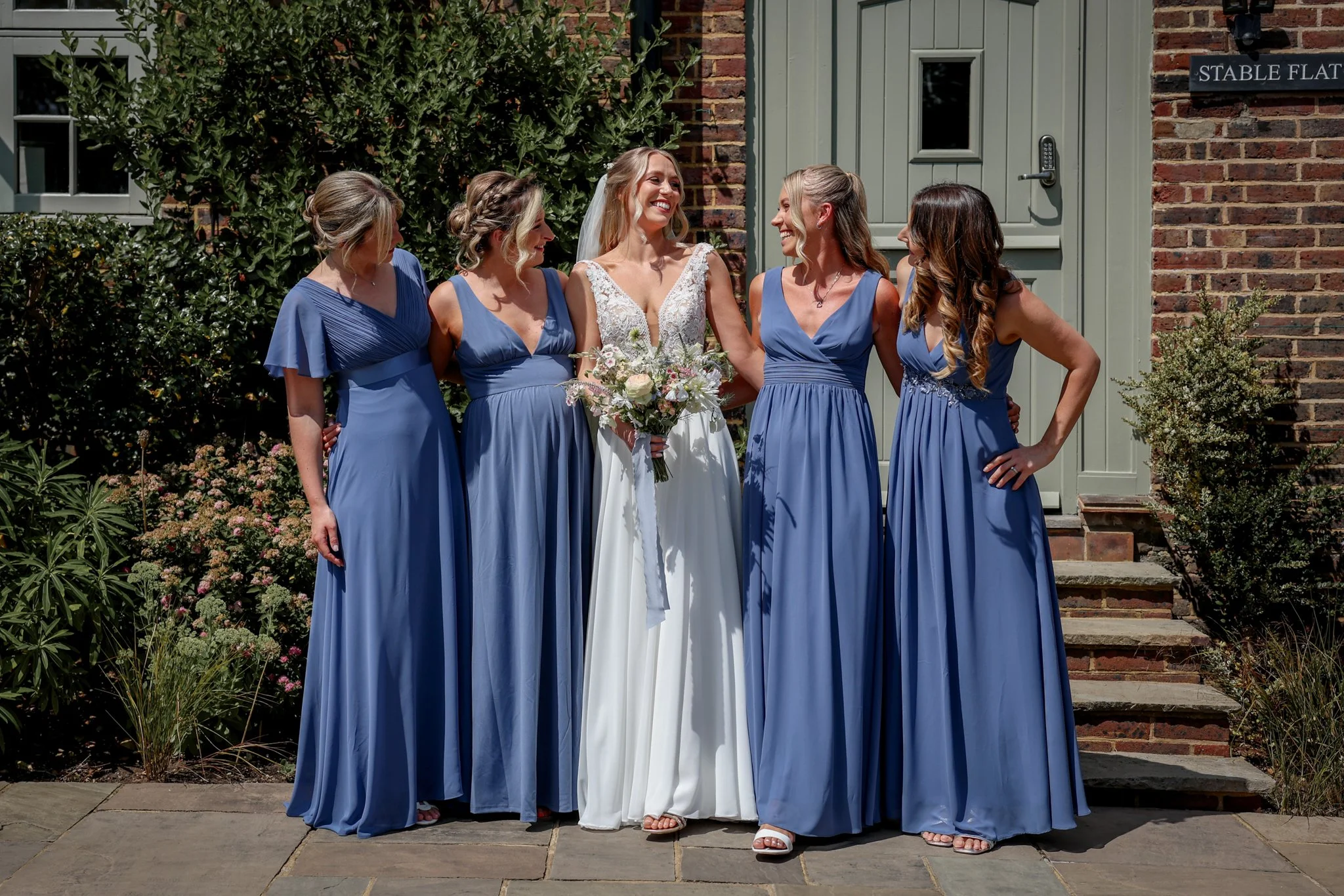 A group of five women, one in a white wedding dress and the other four in matching blue bridesmaid dresses, standing outside in front of a brick building with greenery, smiling and talking together.