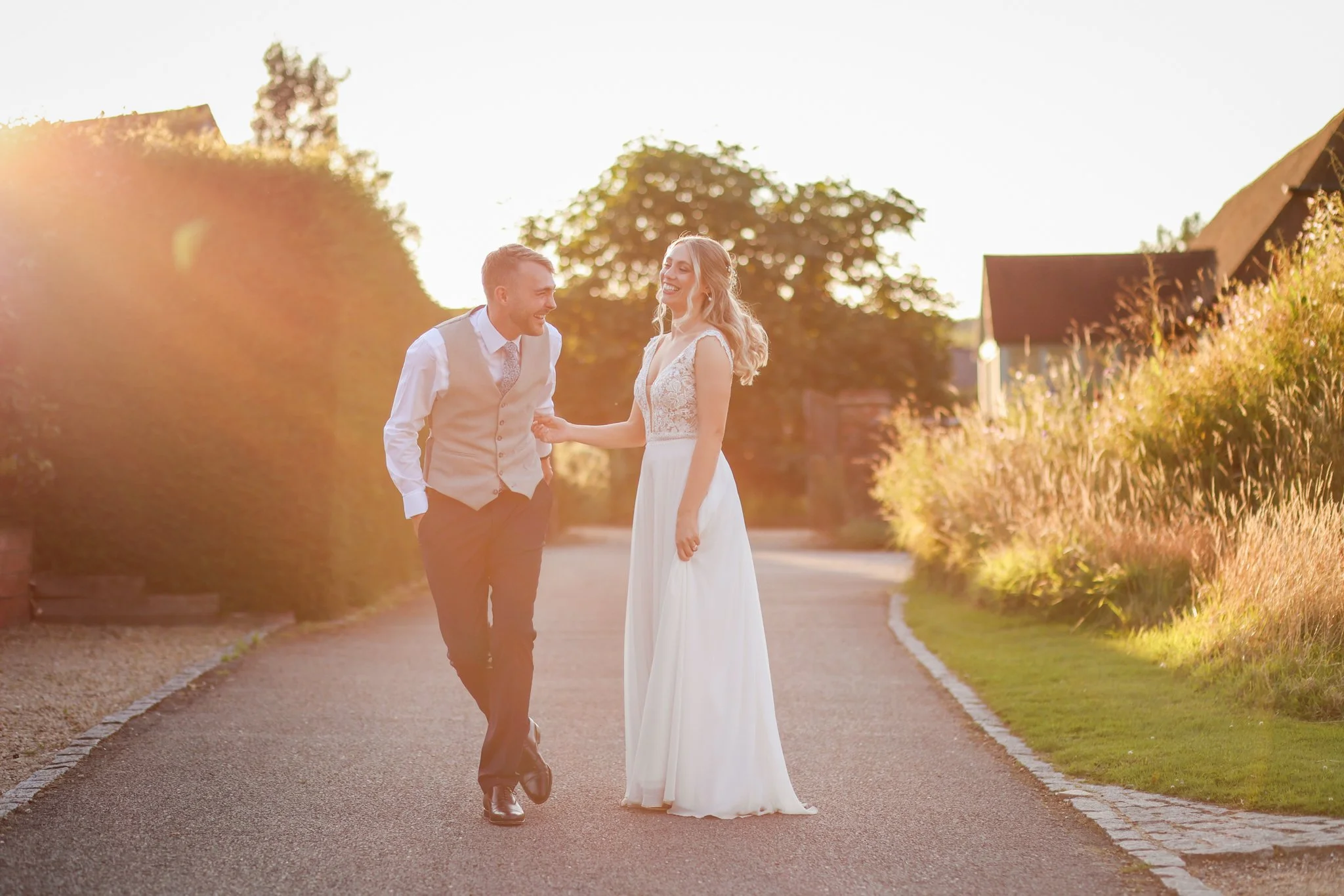 A smiling bride and groom walking hand in hand on a tree-lined pathway during sunset, dressed in wedding attire.