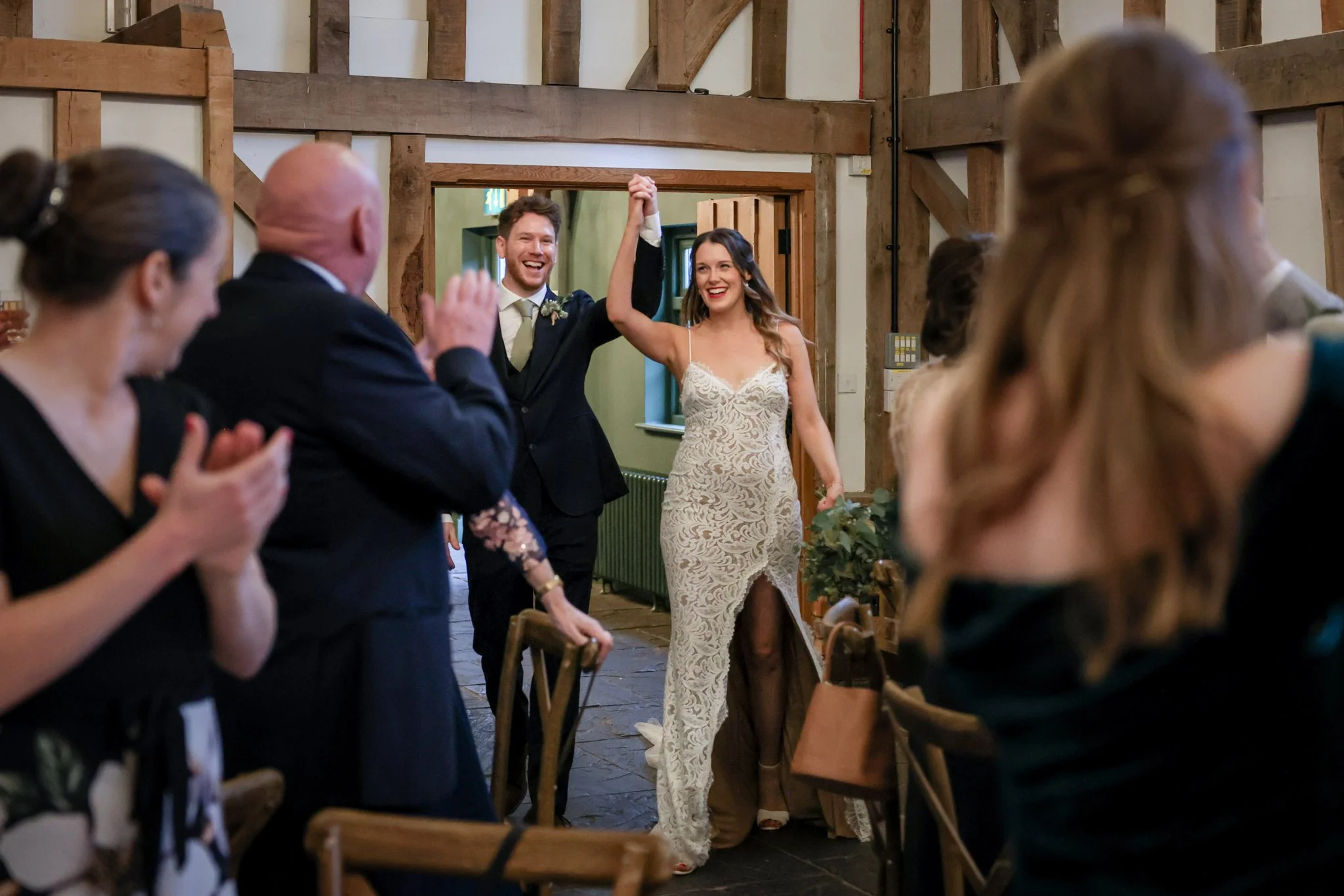 A newlywed couple, a man in a black suit and white shirt and a woman in a lace wedding dress, holding hands and smiling as they walk through a reception with seated guests clapping and celebrating.