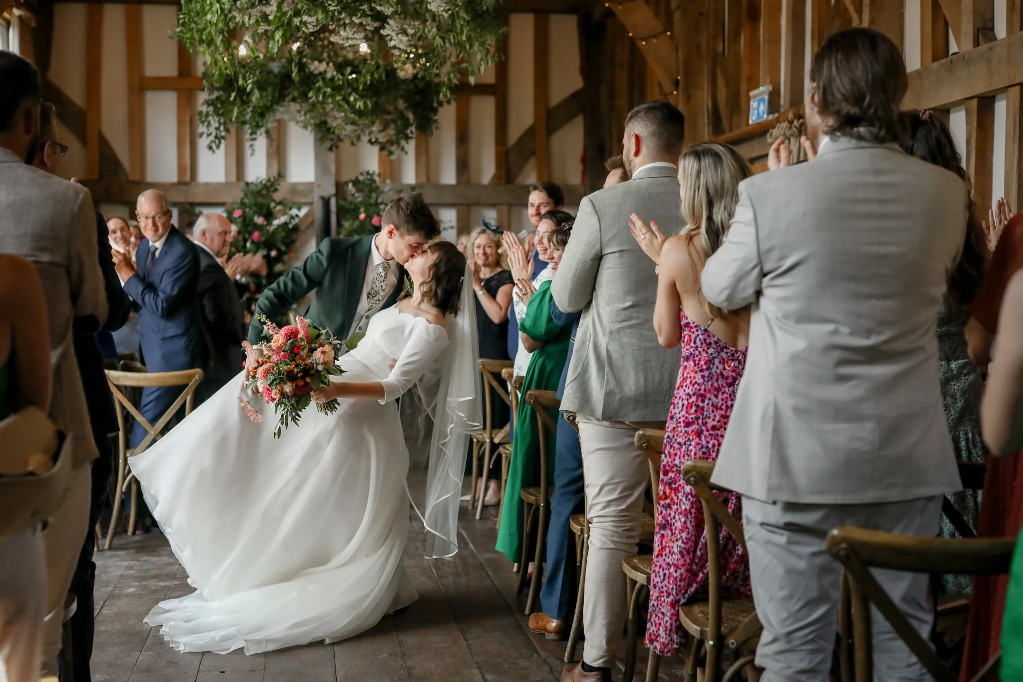 A bride and groom share a kiss at their wedding ceremony surrounded by seated and standing guests clapping and smiling.
