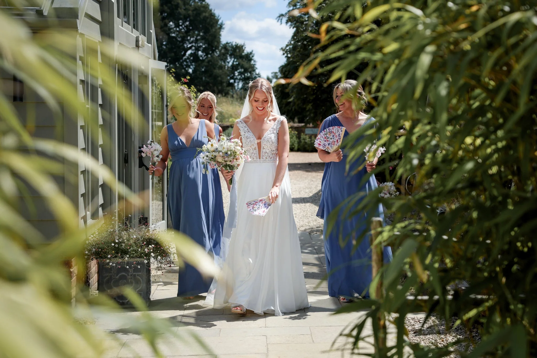 A bride in a white wedding dress smiling while walking outdoors with three women, likely bridesmaids, dressed in blue dresses, holding flowers. They are near a building with greenery and trees in the background, on a sunny day.