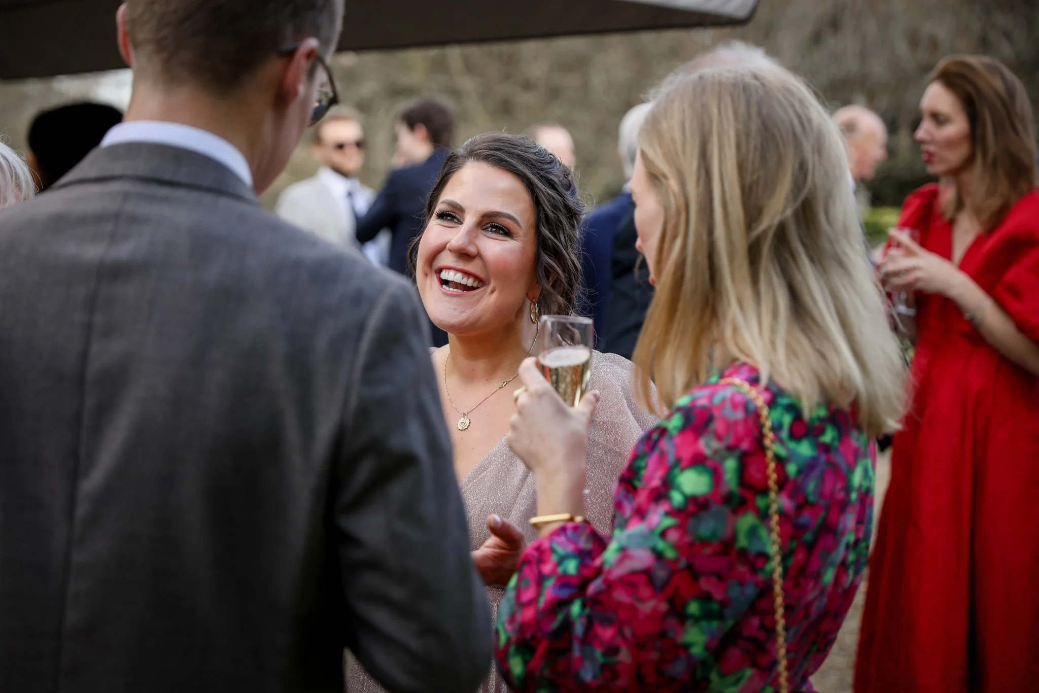 People at a social gathering outdoors, engaging in conversation with smiling faces, with blurred individuals in the background.