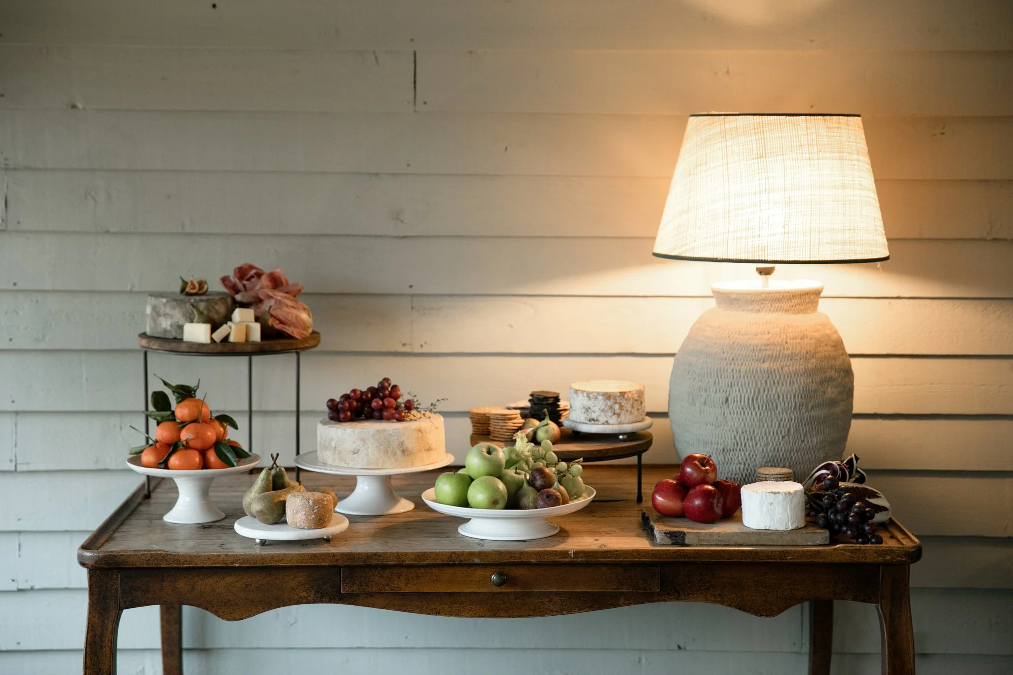 A rustic wooden table displaying various fresh fruits including oranges, apples, pears, grapes, and miscellaneous produce, with a table lamp illuminating the scene against a white wooden wall background.