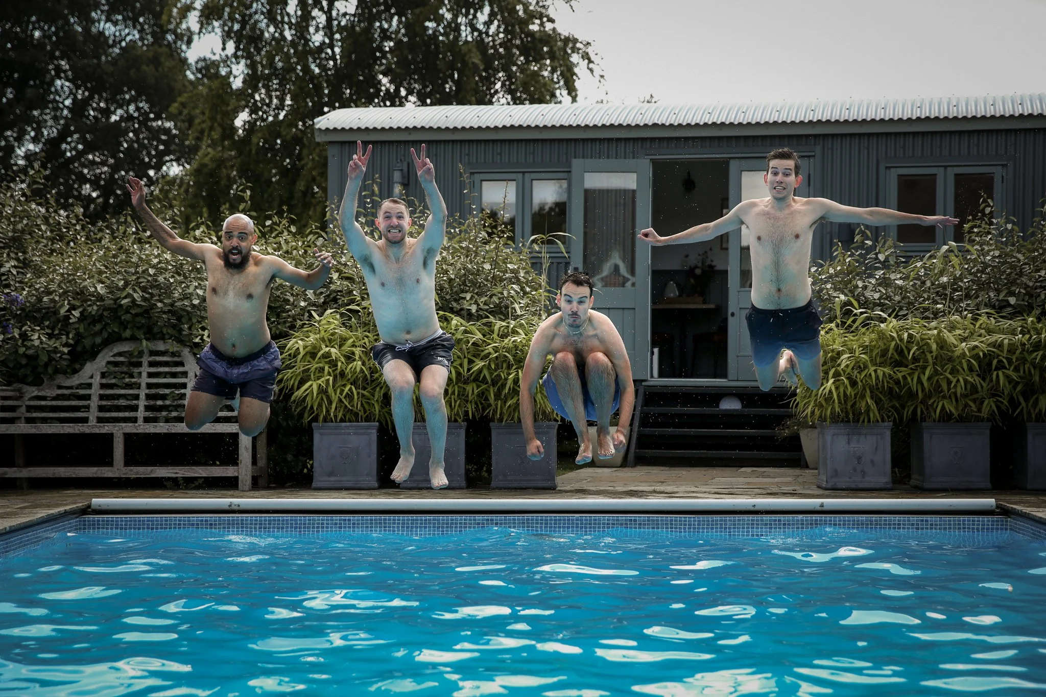 Four men jumping into a swimming pool from a wooden deck, with one sitting and three in mid-air. They are outdoors, next to a small building, and surrounded by greenery.