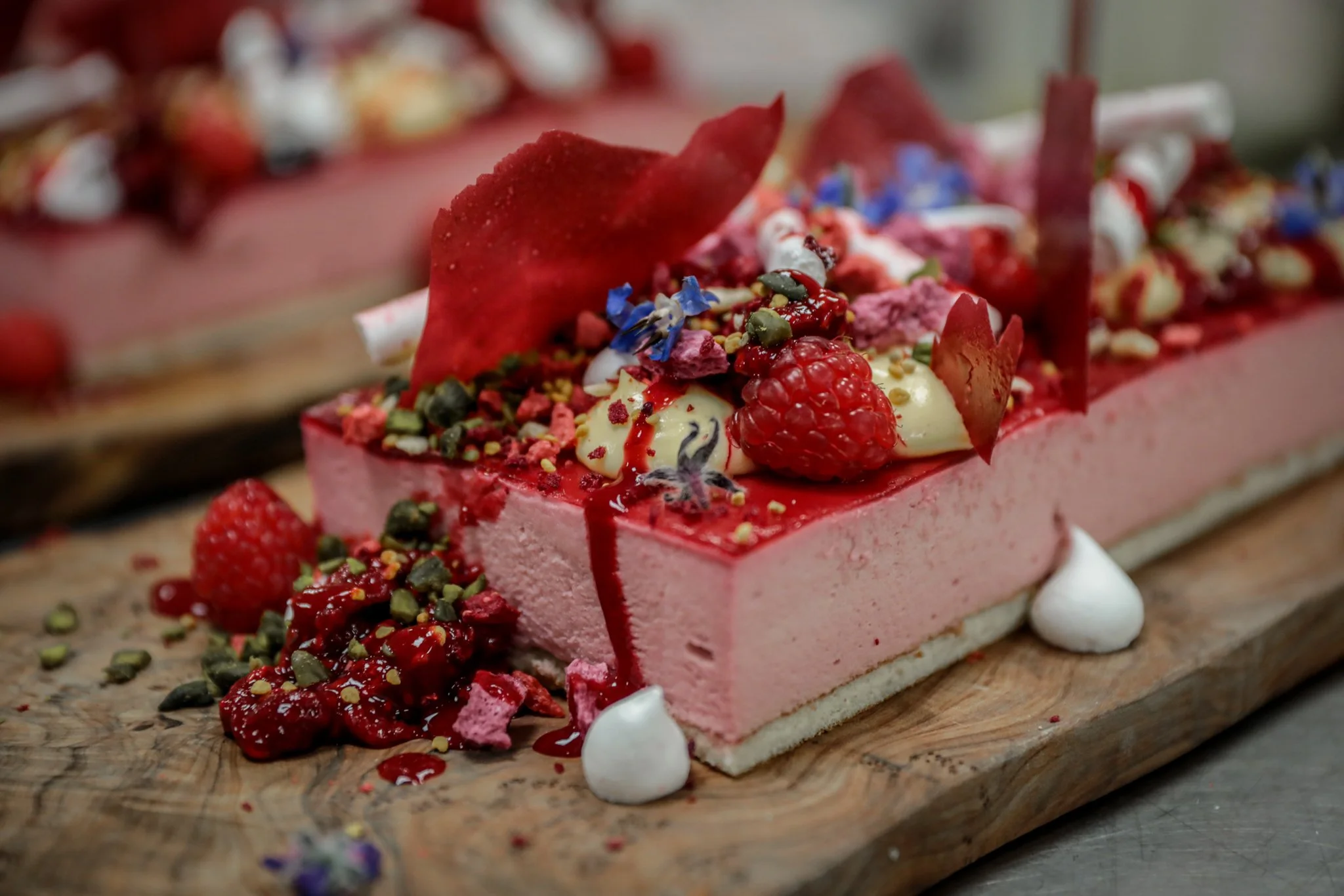 Close-up of a rectangular pink layered cake decorated with red fruit, edible flowers, seeds, and drizzled with red sauce, placed on a wooden board.