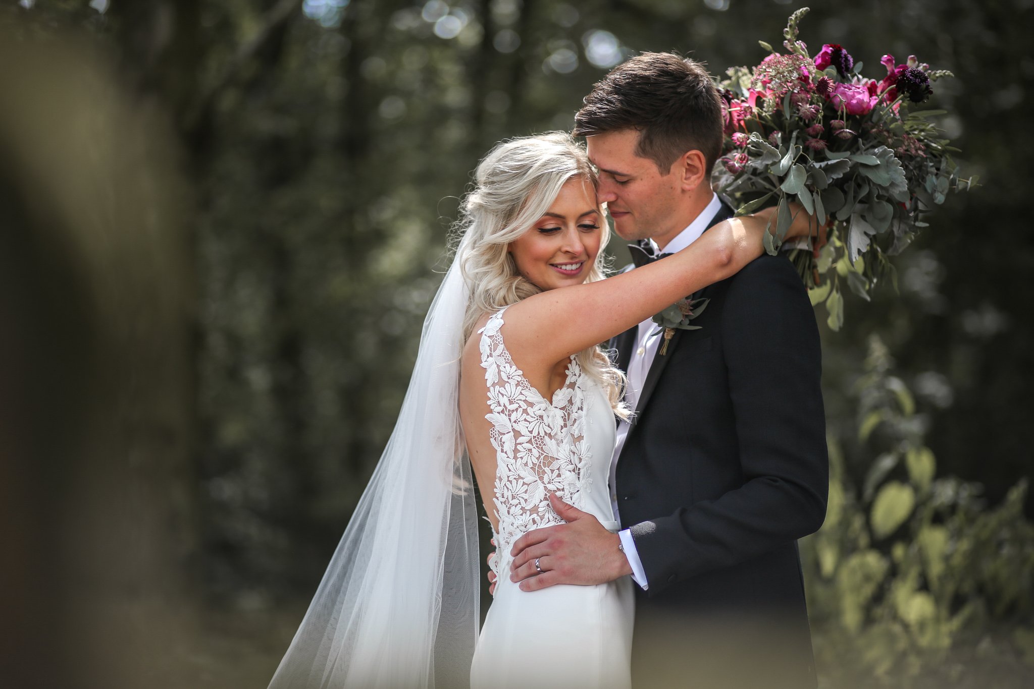 A bride and groom sharing an intimate moment during their outdoor wedding, with the bride holding a large bouquet of pink and purple flowers over her shoulder.