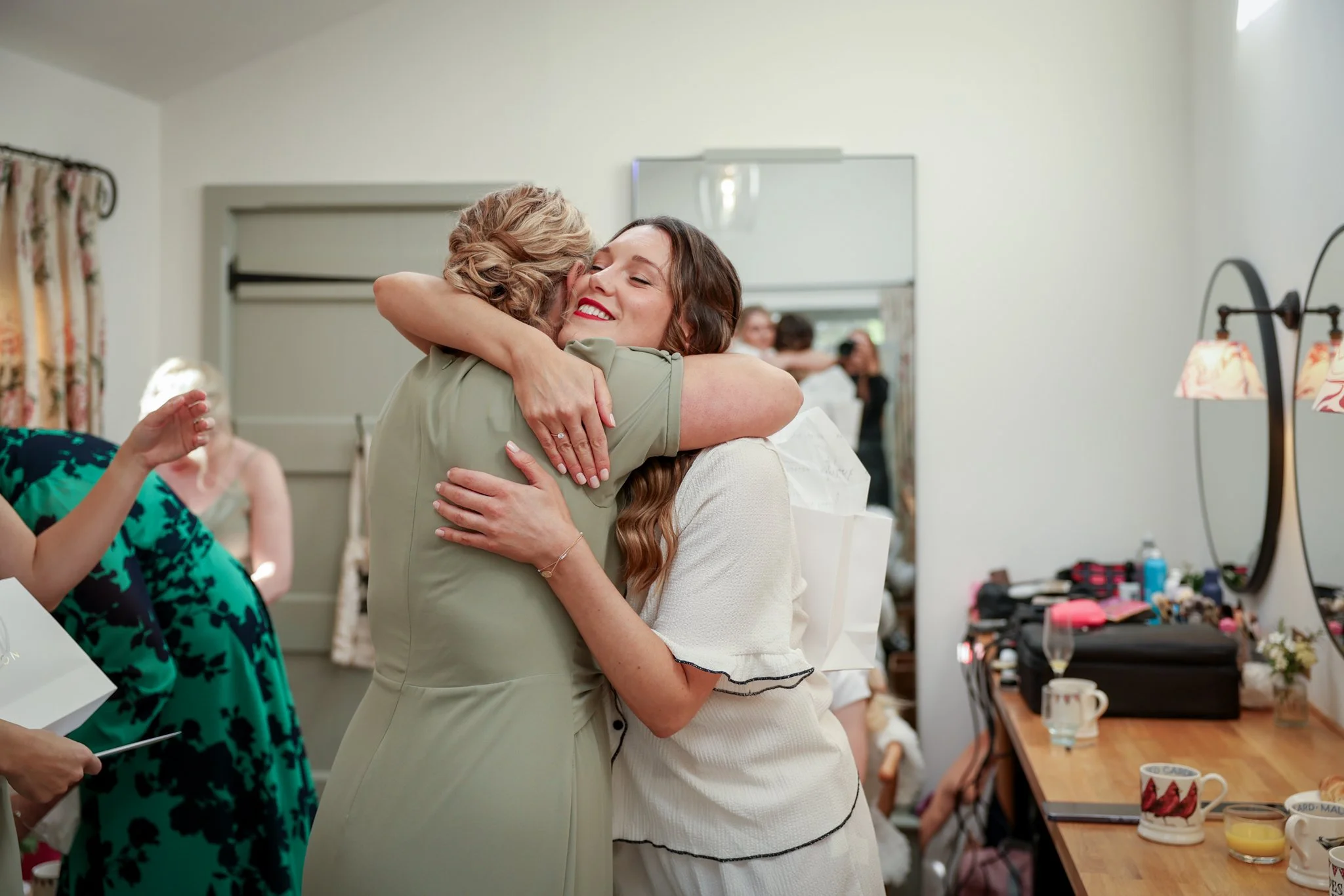 Two women are hugging and smiling in a cheerful setting, possibly a celebration or gathering at a home.
