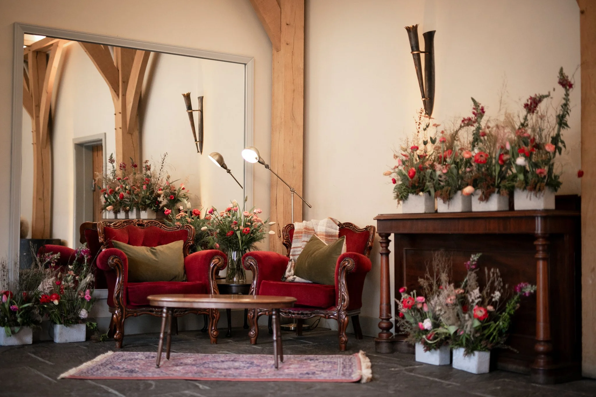 A cozy living room decorated with pink and red flowers in white planters, red velvet sofas, green cushions, a wooden coffee table, a large mirror, and wall-mounted torches on a neutral wall.