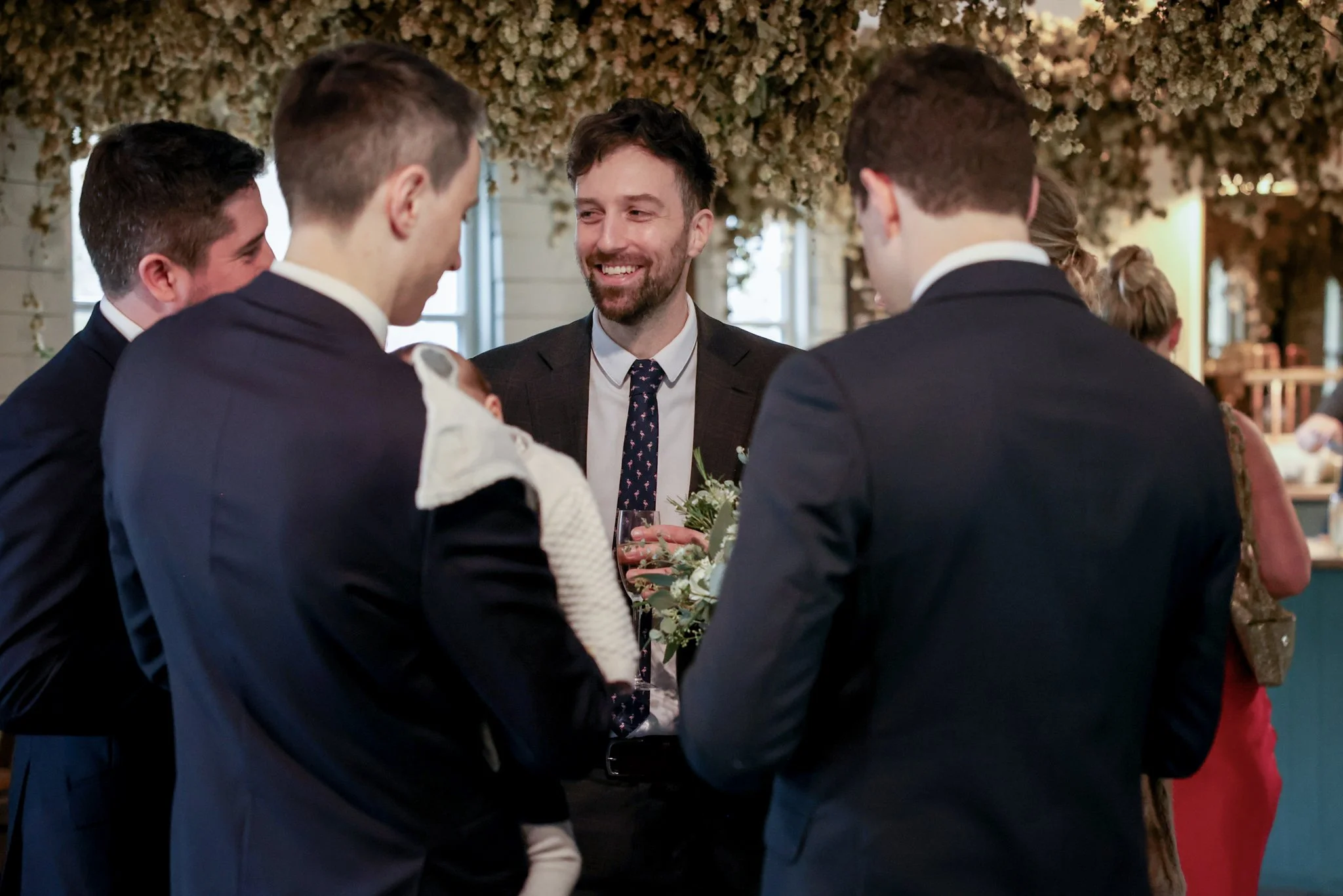 A group of five people dressed in formal attire, gathered and smiling at each other in a decorated indoor setting with floral arrangements overhead.