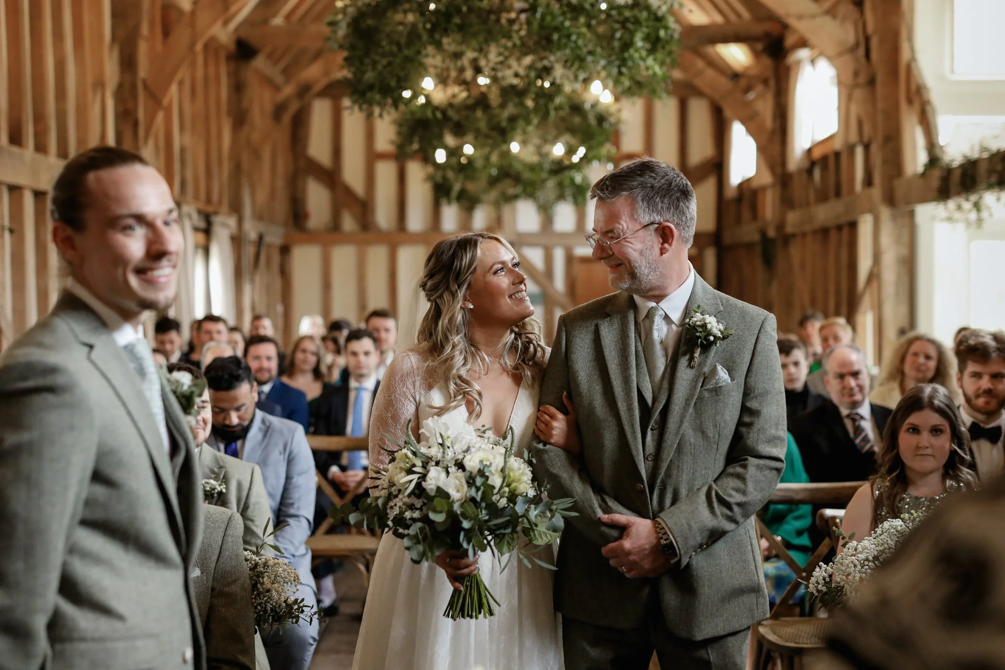 A bride and an older man, likely her father, smiling and holding hands at a wedding ceremony in a rustic barn with wooden beams and greenery hanging from the ceiling. The bride holds a bouquet of white flowers
