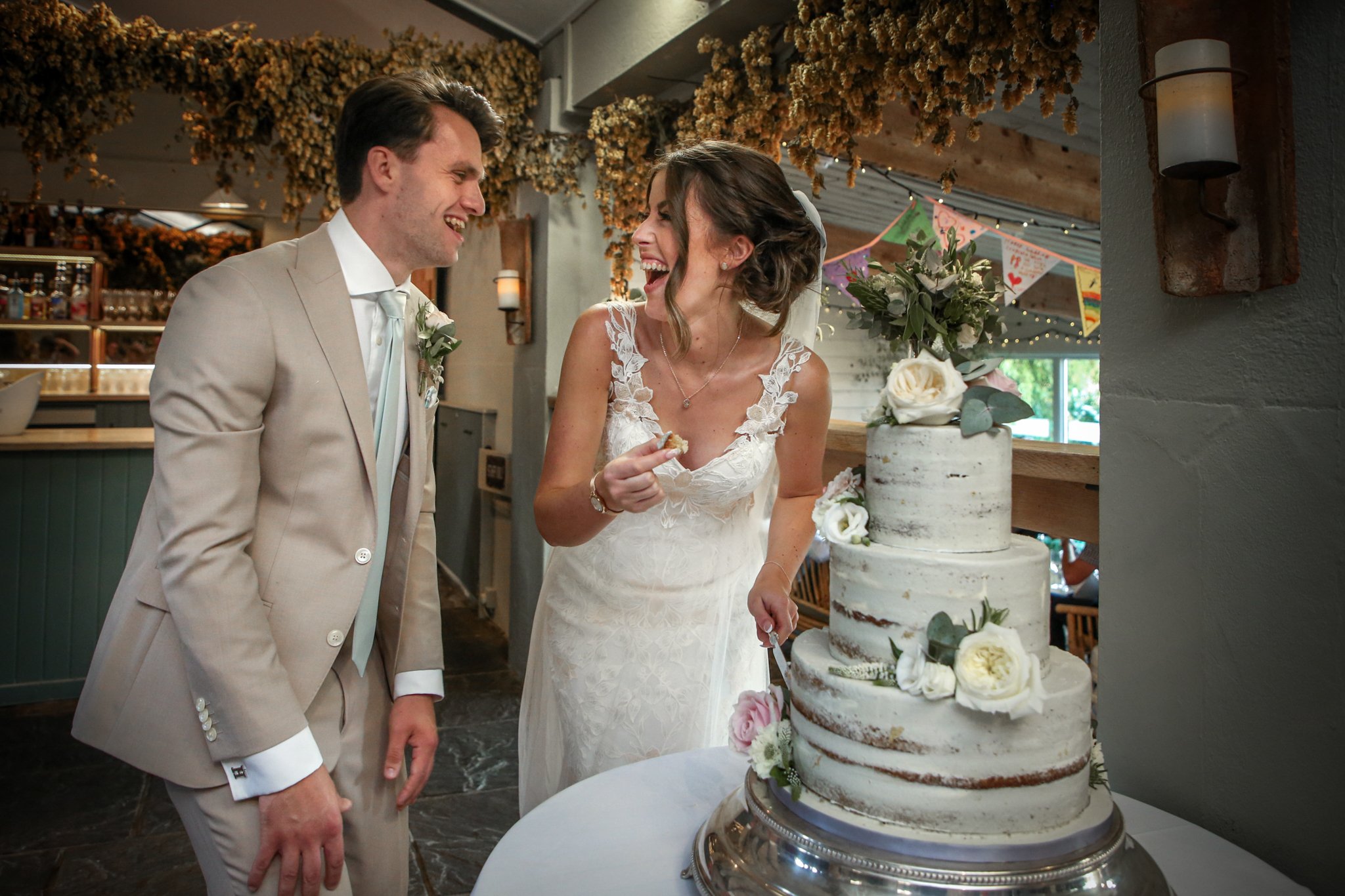 A bride and groom laughing and smiling at a wedding cake, decorated with white and pink flowers, in a decorated venue.