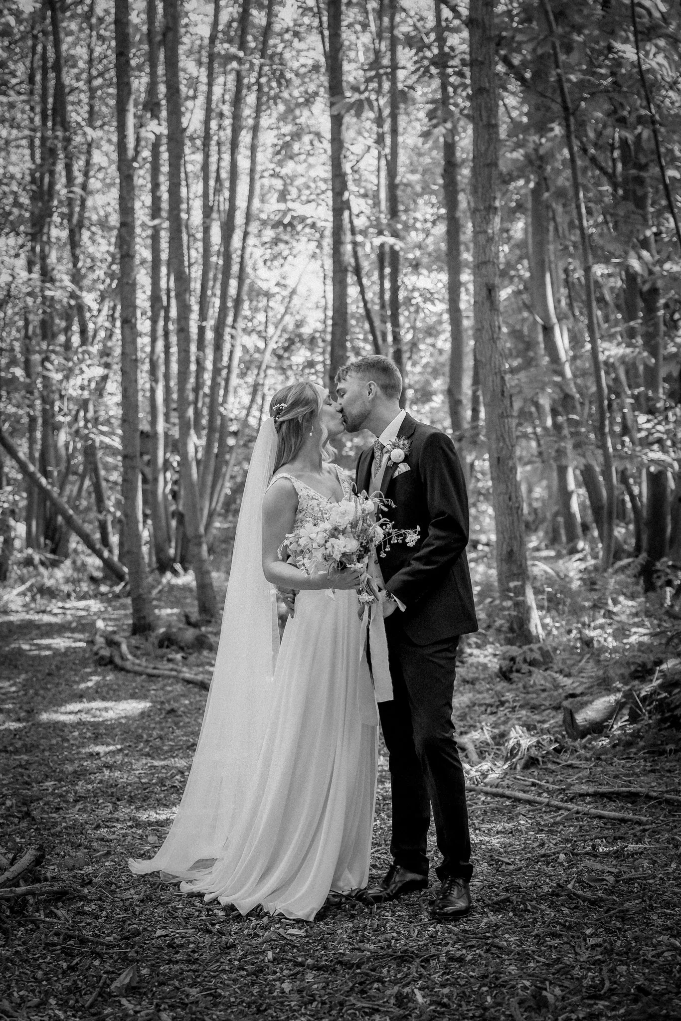 Black and white photo of a bride and groom standing close together in a wooded area, sharing a kiss. The bride is in a long wedding dress holding a bouquet, and the groom is in a suit with a tie and boutonniere.