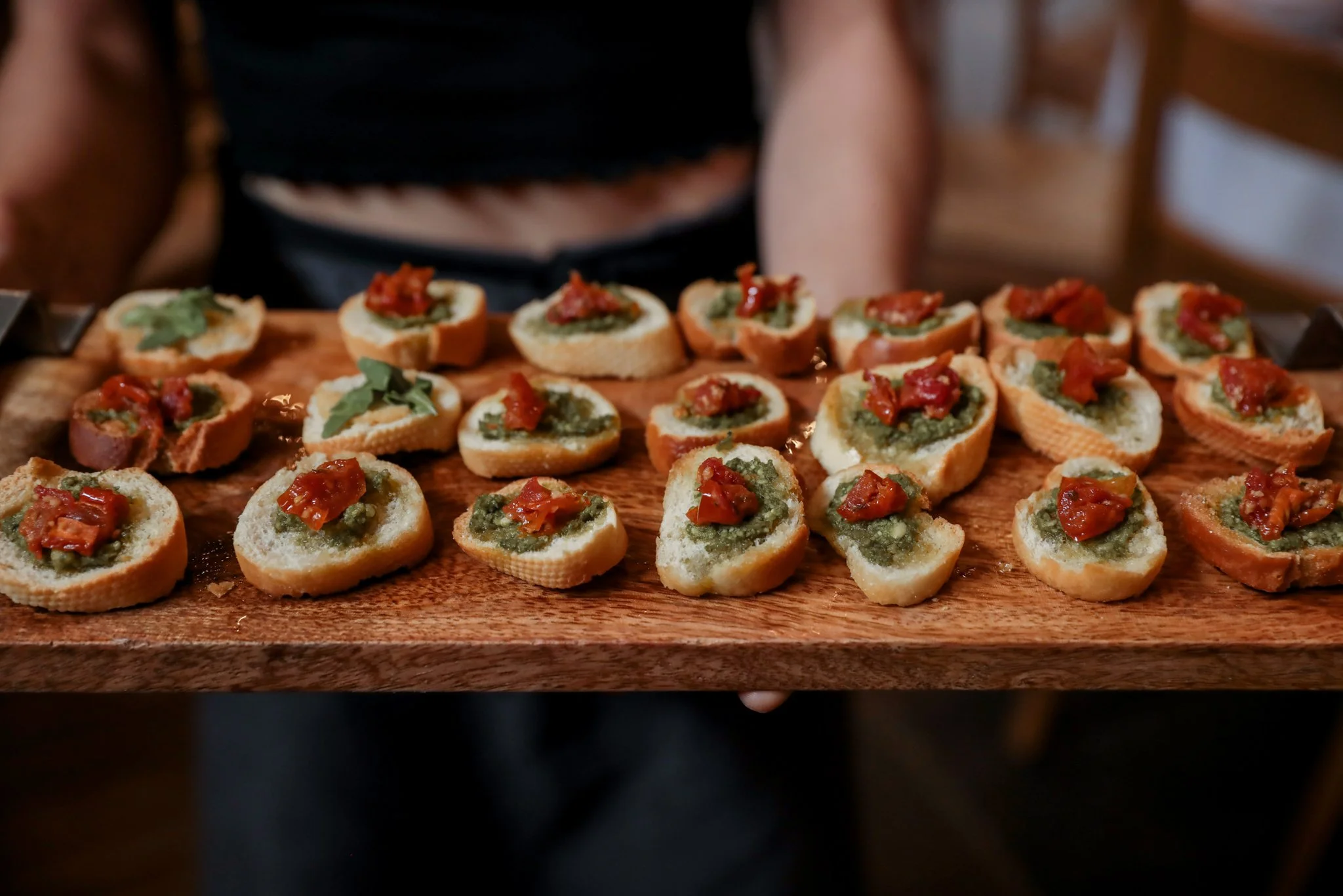 Video still of a person presenting a wooden board with arranged bread slices topped with green spread, tomato, and herbs.