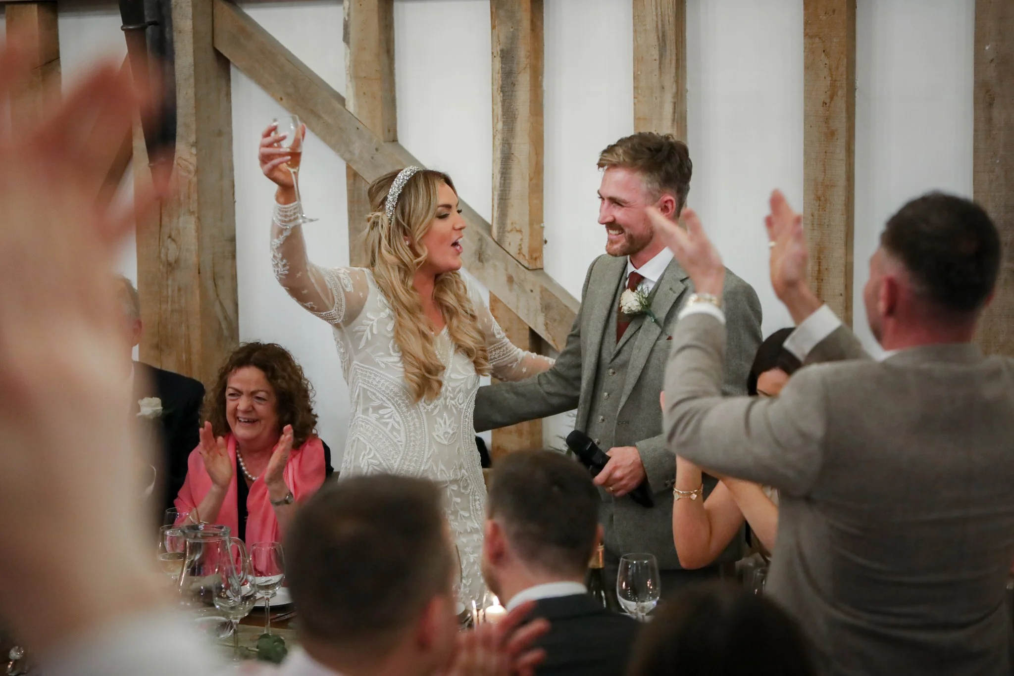 A bride and groom at their wedding reception, smiling and holding hands, surrounded by guests who are clapping and celebrating.
