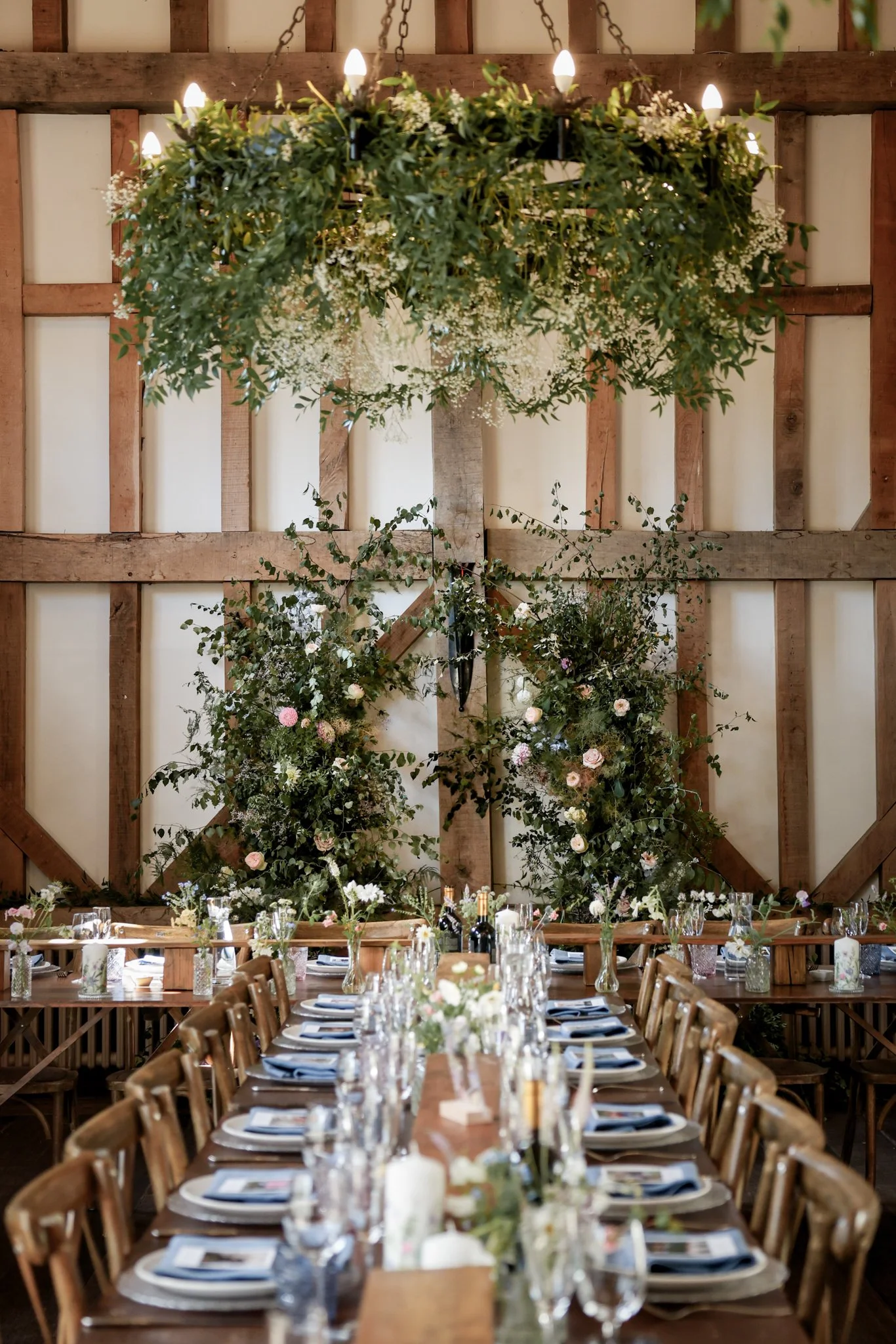 A rustic banquet hall with a long wooden table set for a celebration. The table has glassware, plates, and napkins, and is decorated with floral arrangements. A large floral and greenery hanging chandelier with candles and lights hangs above the tabl