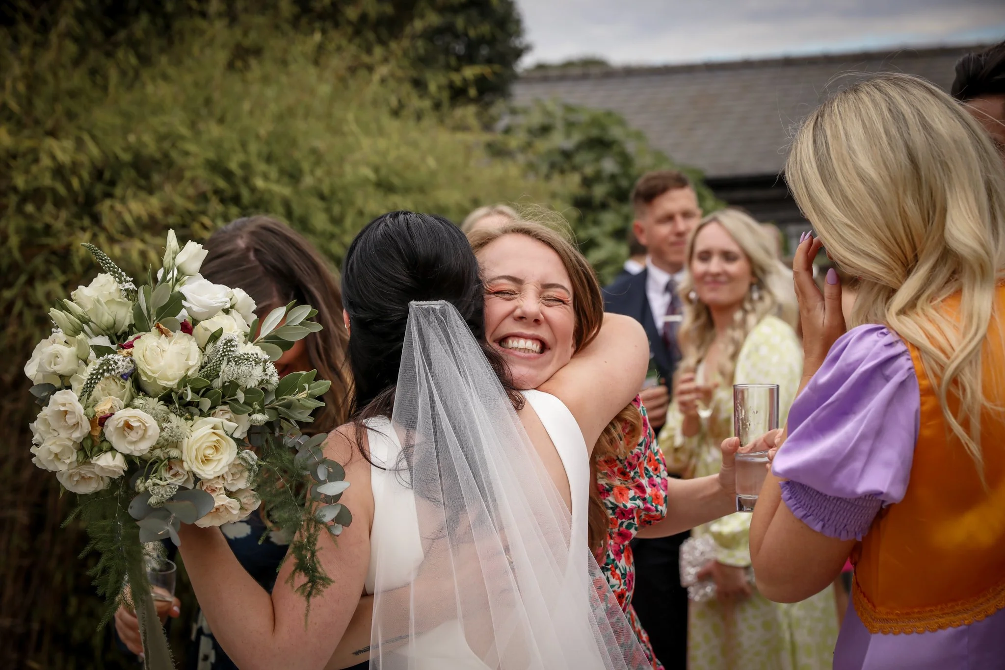 A woman in a white dress with a veil hugs another woman during a celebration, with a bouquet of white roses and greenery nearby, and other guests in the background outdoors.