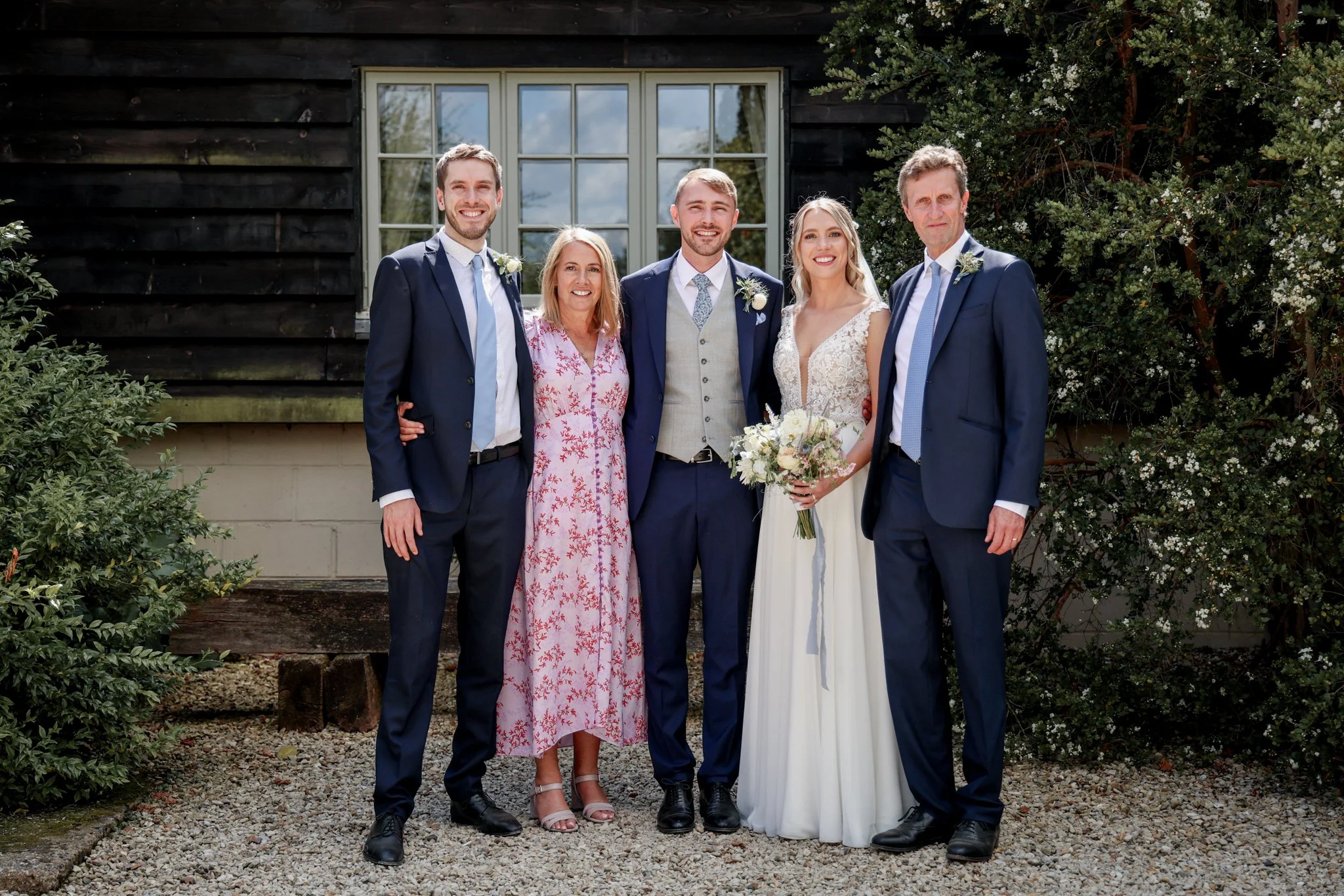 Group of six people, three men and three women, standing close together outdoors in front of a black wooden building with a large window, during a daytime celebration or wedding. They are dressed in formal attire, with the women wearing dresses and t