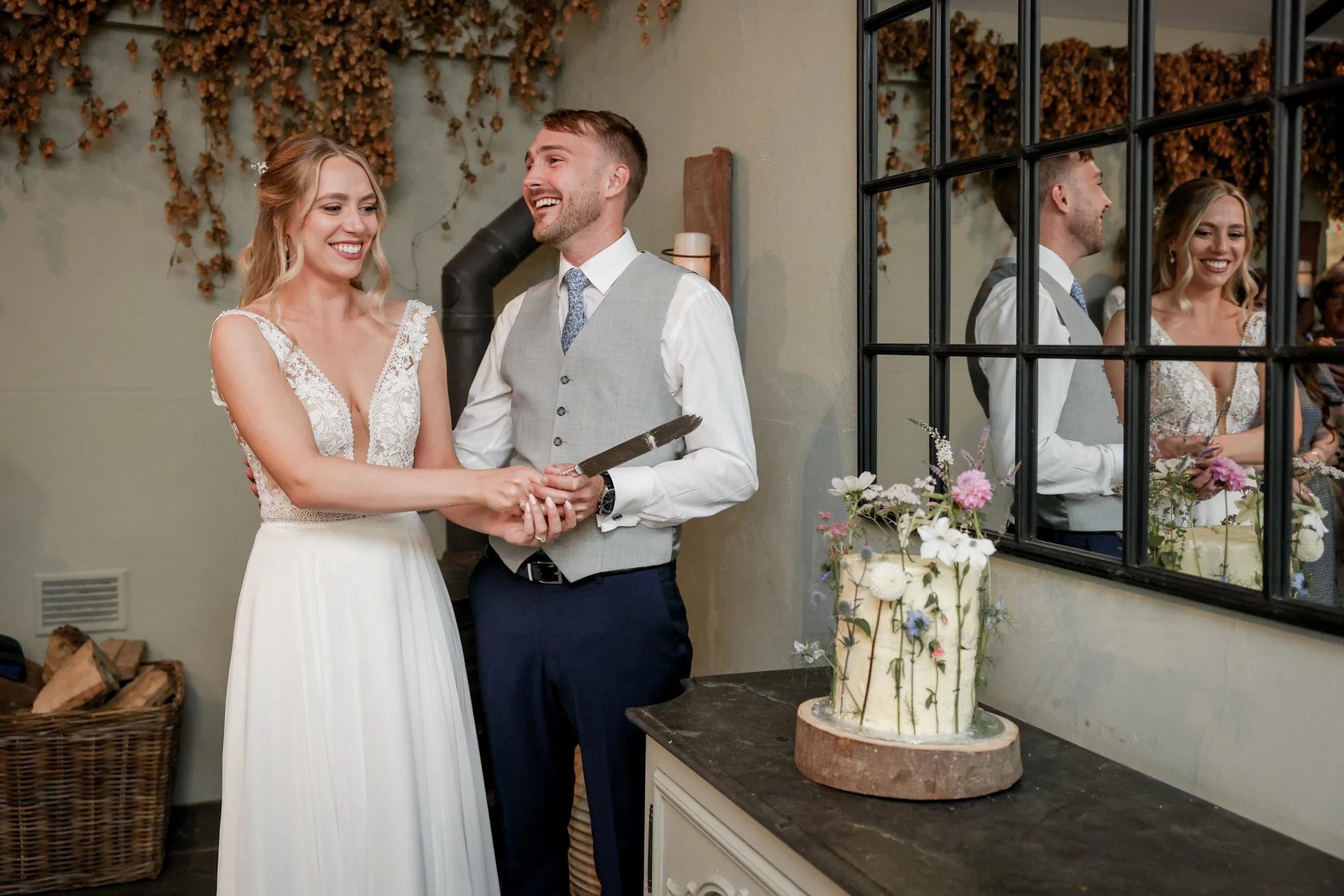 A couple at their wedding ceremony, with the bride in a white lace dress and the groom in a gray vest and tie, holding hands and smiling at each other by a wedding cake with flowers on the table.