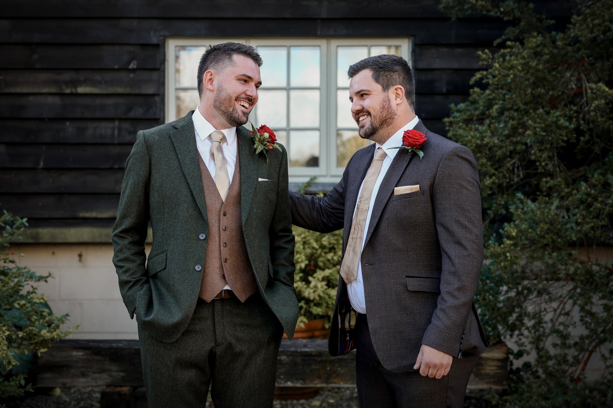 Two men in suits are talking and smiling outside in front of a black wooden building with a window; they are wearing boutonnieres with red roses.