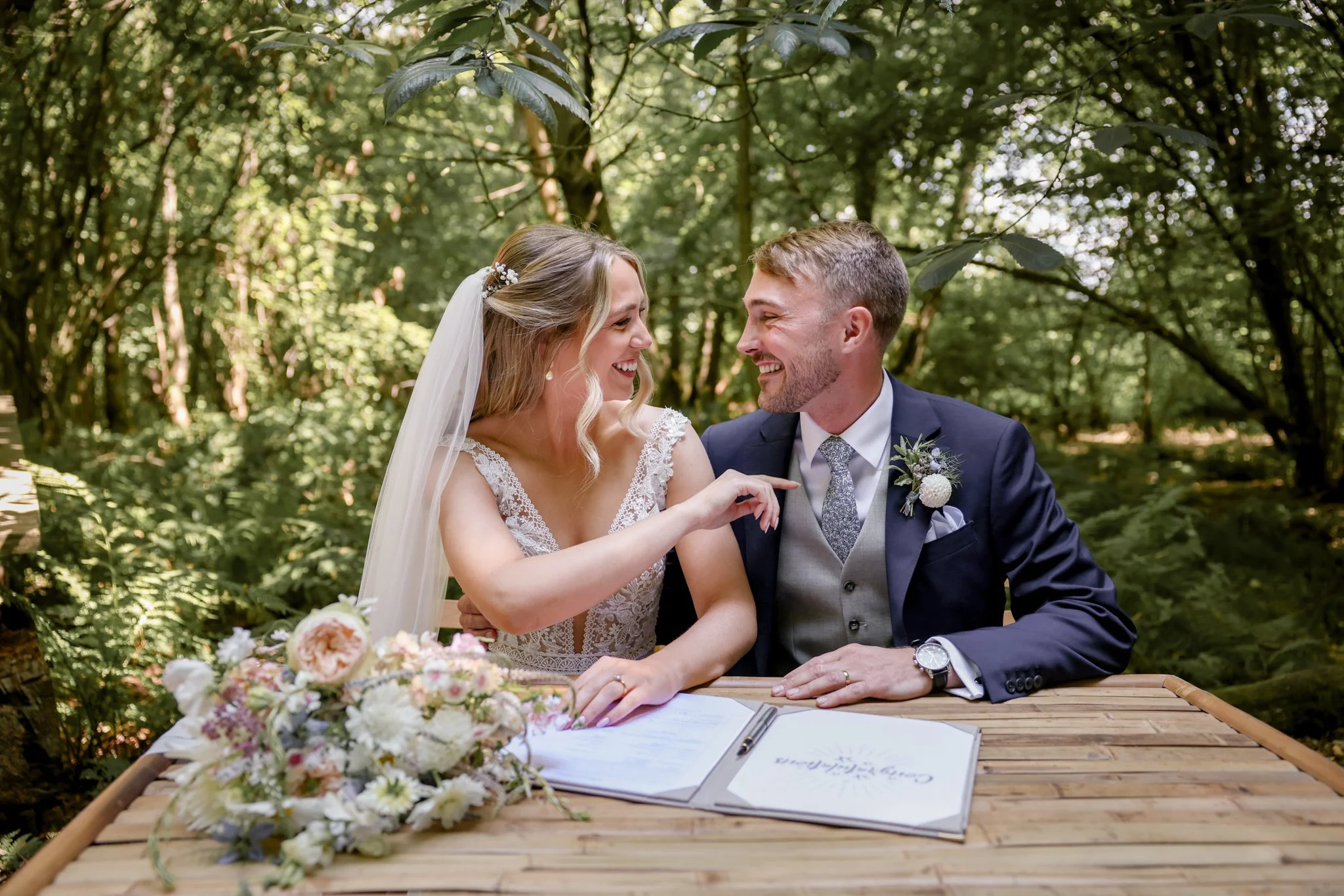 A bride and groom sit at a wooden table outdoors in a forest during a wedding ceremony, smiling at each other with a bouquet of flowers in front of them.