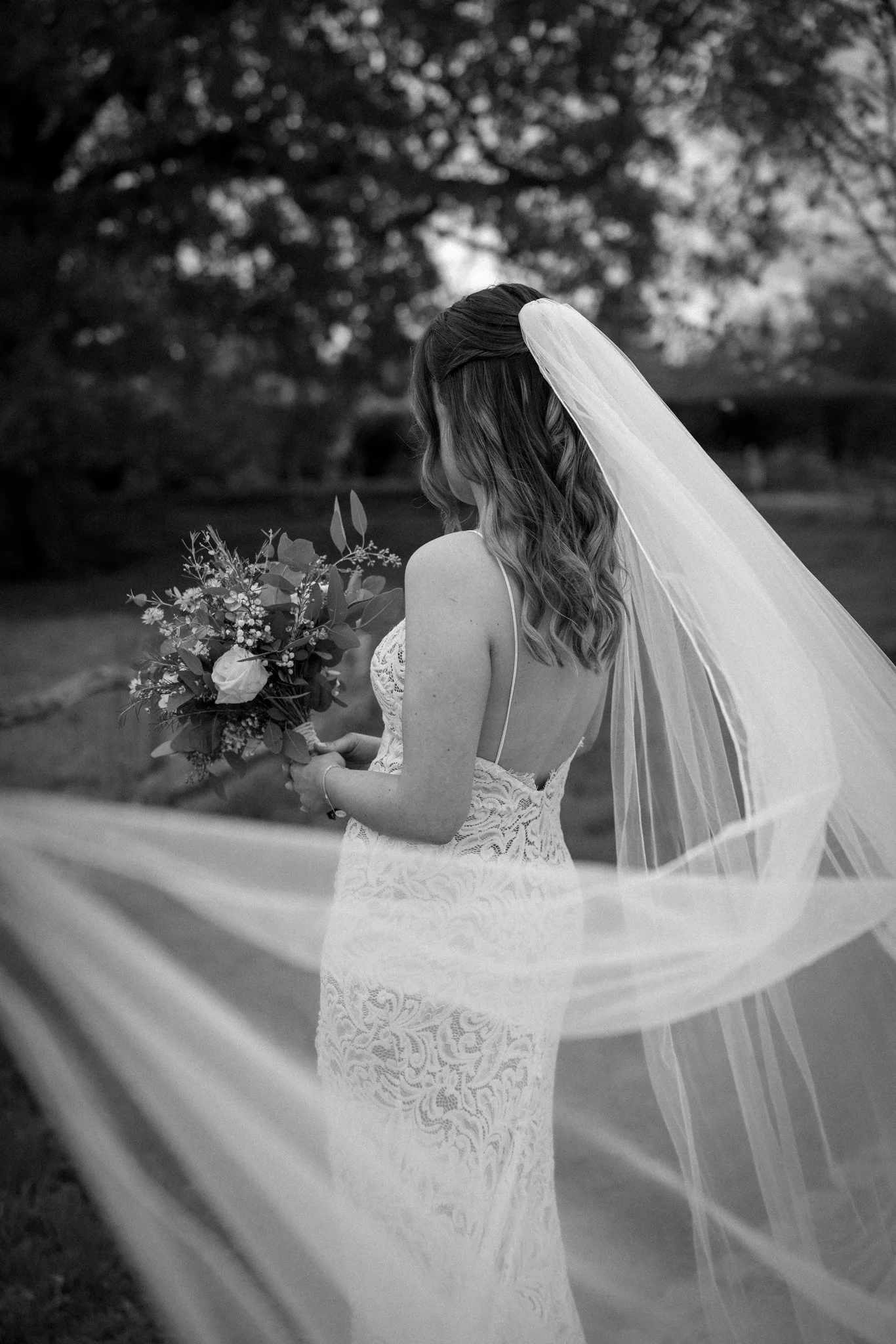 A black and white photo of a bride holding a bouquet of flowers, with her face turned away, wearing a lace wedding dress and a long veil, outdoors in a natural setting.