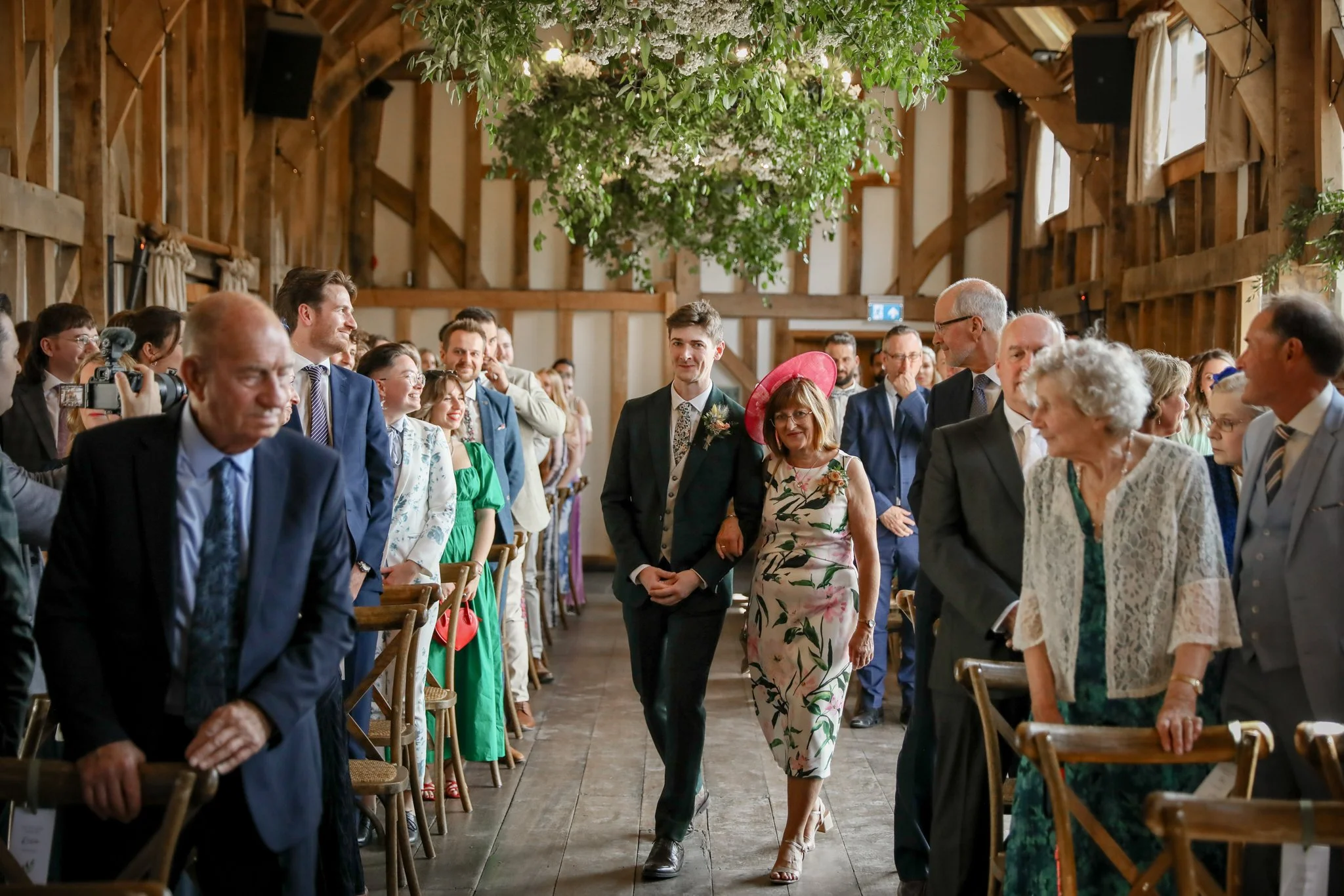 A group of wedding guests seated and standing in a rustic barn with wooden walls and green foliage hanging from the ceiling, watching a couple walk down the aisle during a wedding ceremony.