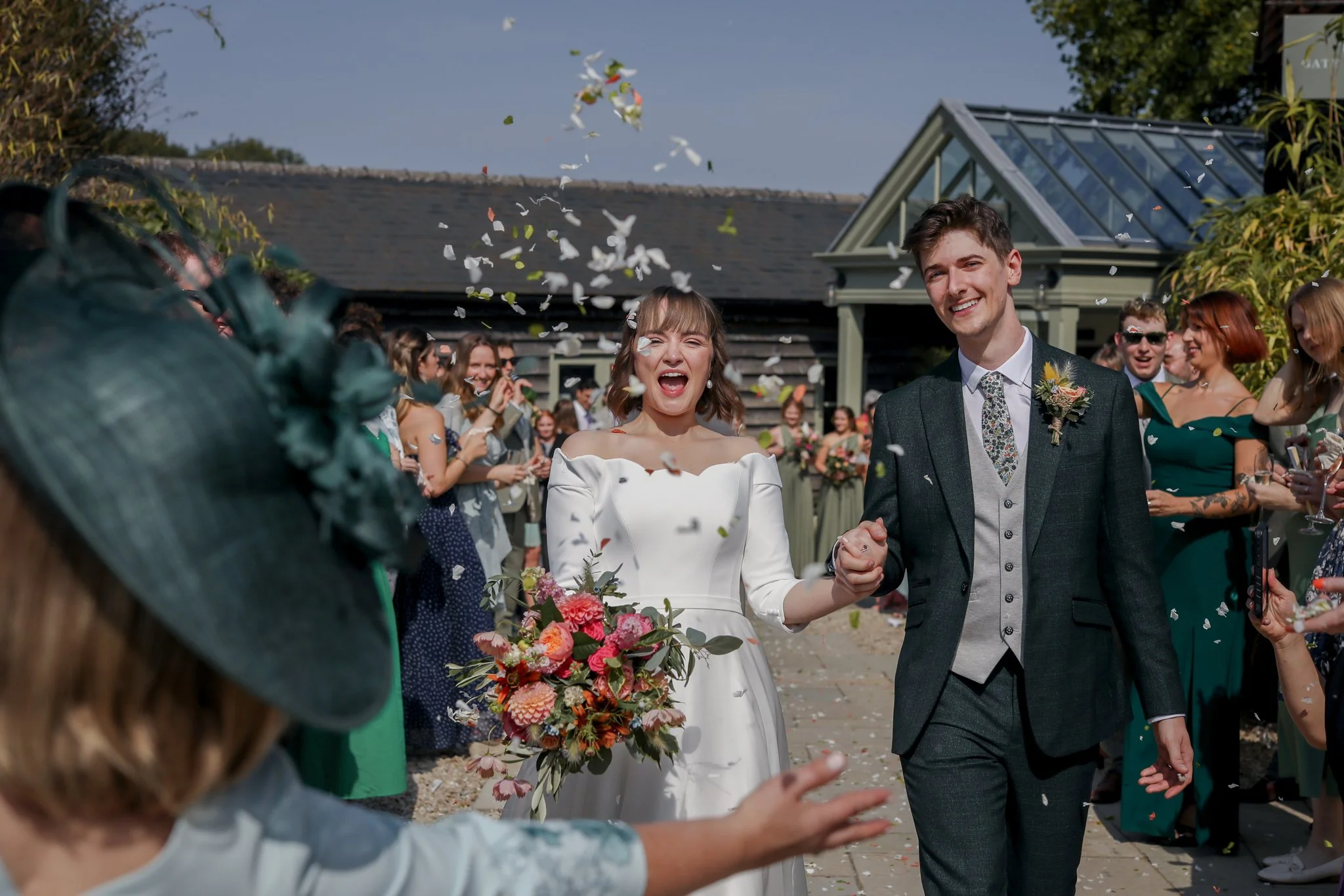 A bride and groom are smiling and holding hands at their outdoor wedding reception, surrounded by guests, with flower petals in the air.