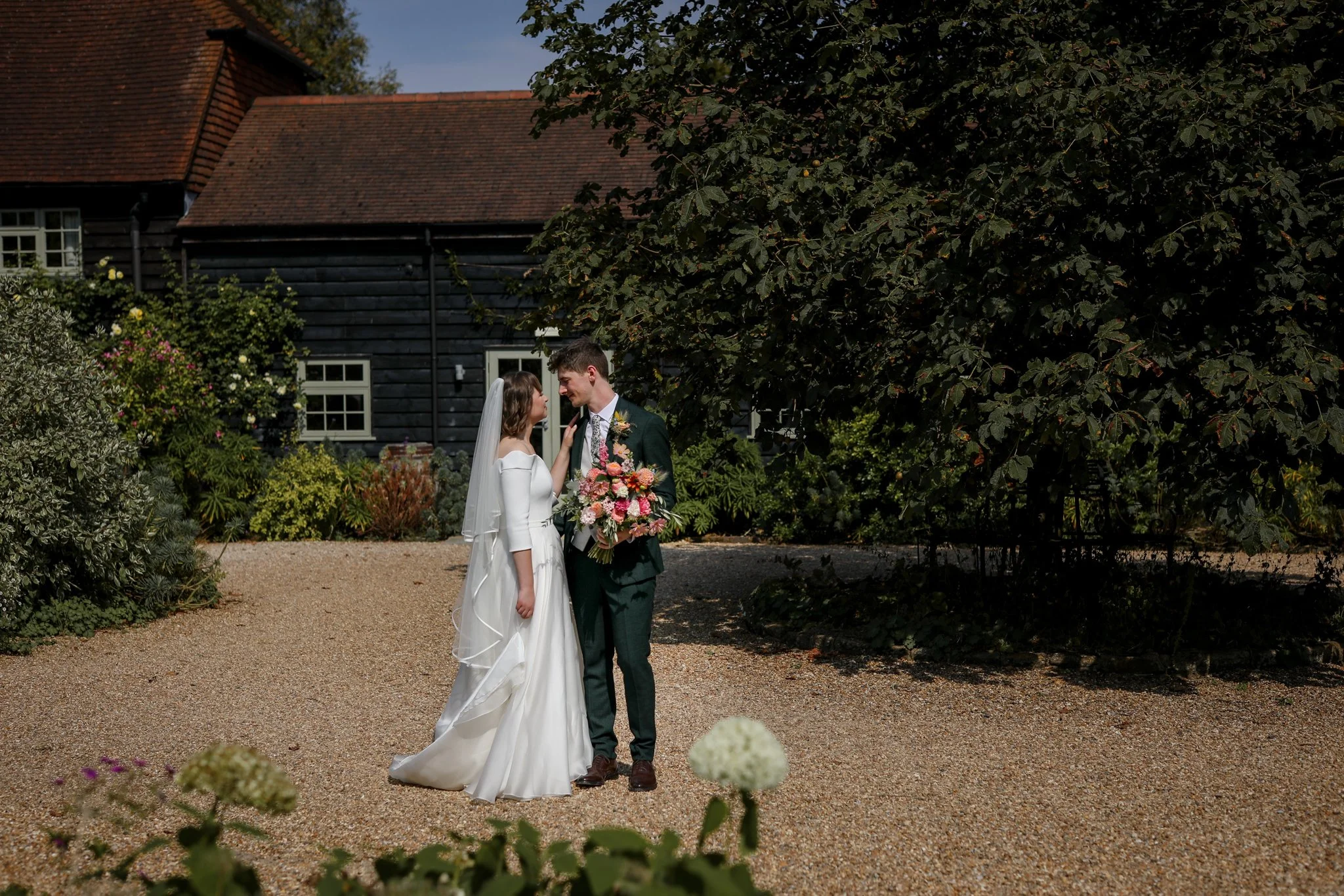 A bride and groom are standing close together in a garden, smiling at each other. The bride is wearing a white wedding dress and veil, holding a large bouquet of pink and peach flowers. The groom is dressed in a green suit with a floral tie. They are